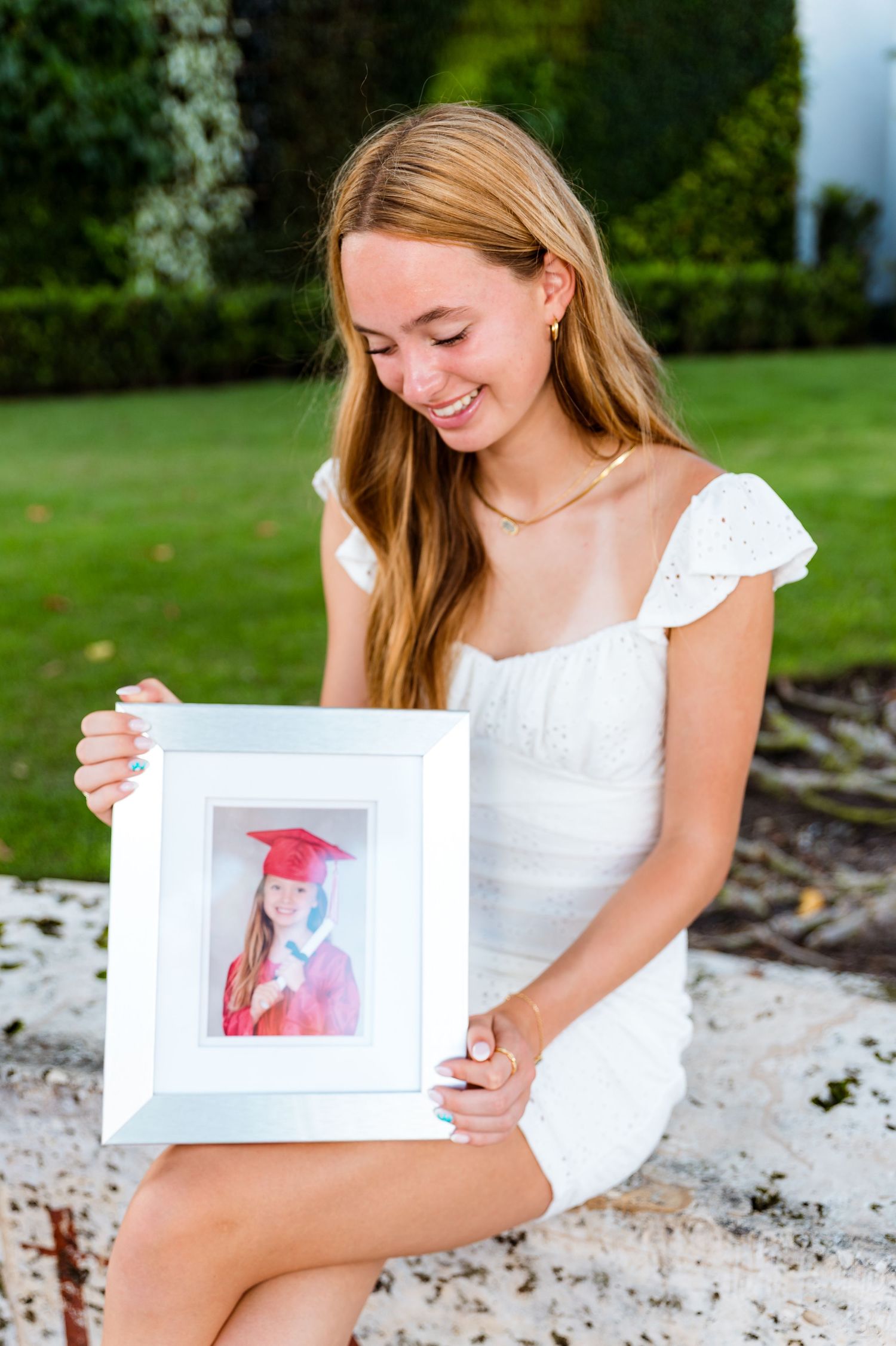 A person in a white dress sits outdoors holding a framed graduation photo against a garden background.