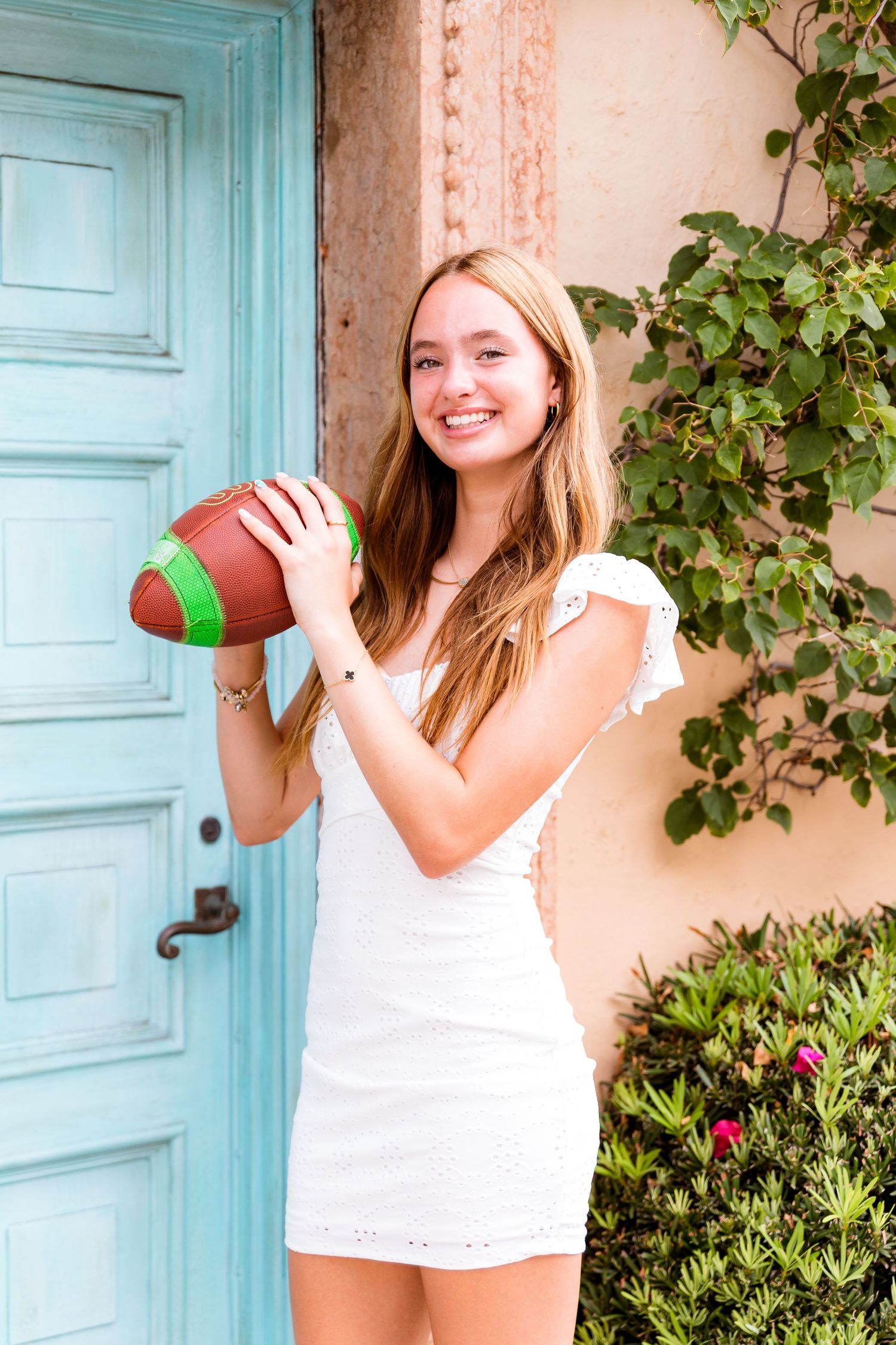 Someone poses with a football while wearing a white dress against a turquoise door and coral wall.