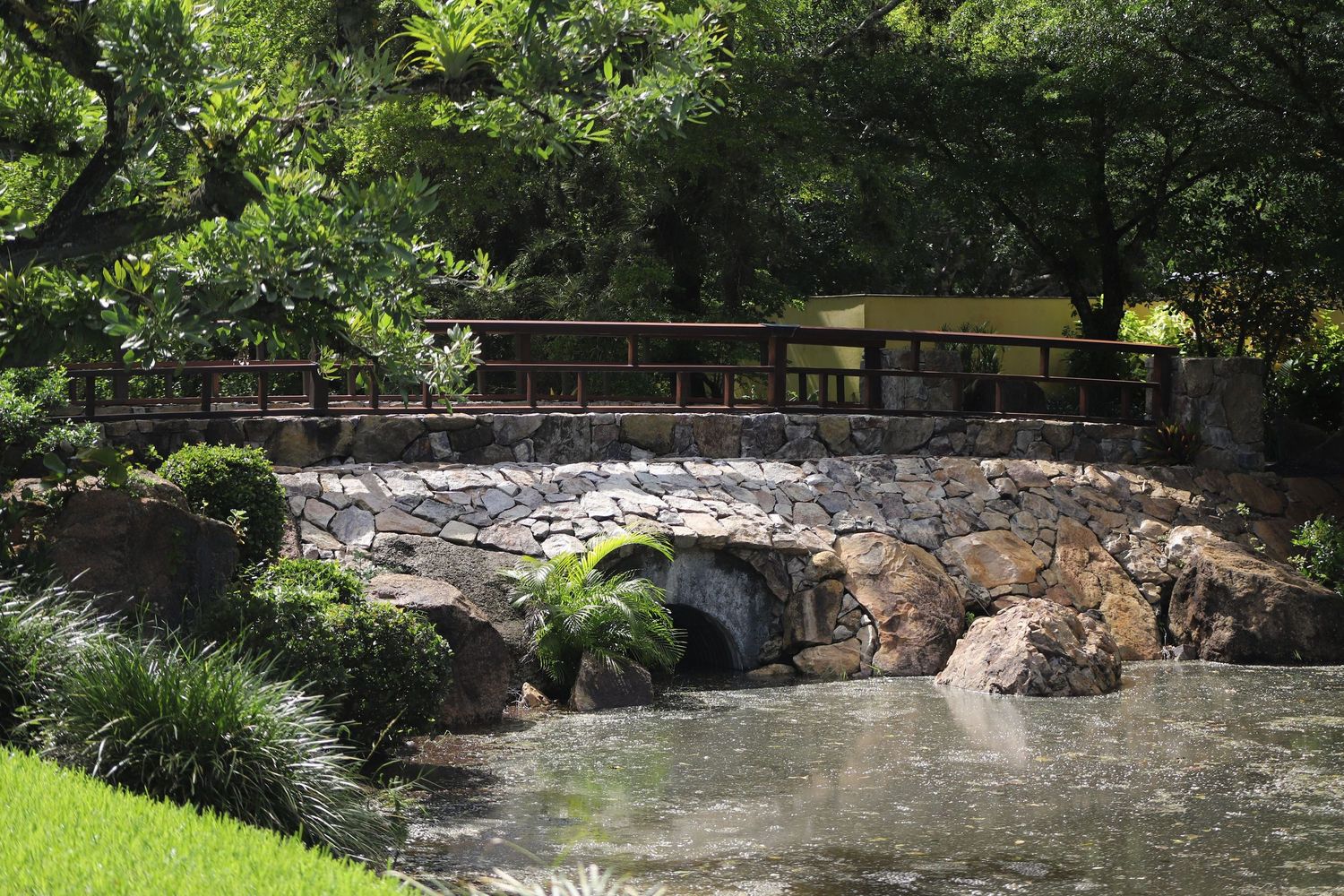 A rustic wooden bridge spans over a rocky stream surrounded by lush greenery and landscaping in a peaceful garden setting.