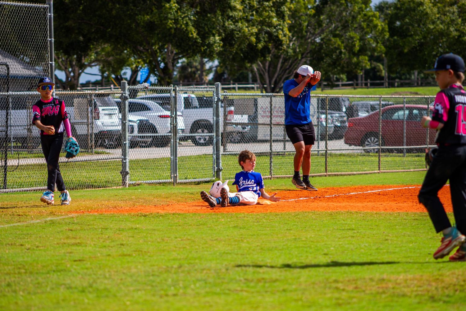 Baseball players practicing fielding drills on a sunny day at a grass field with fences and trees in the background.
