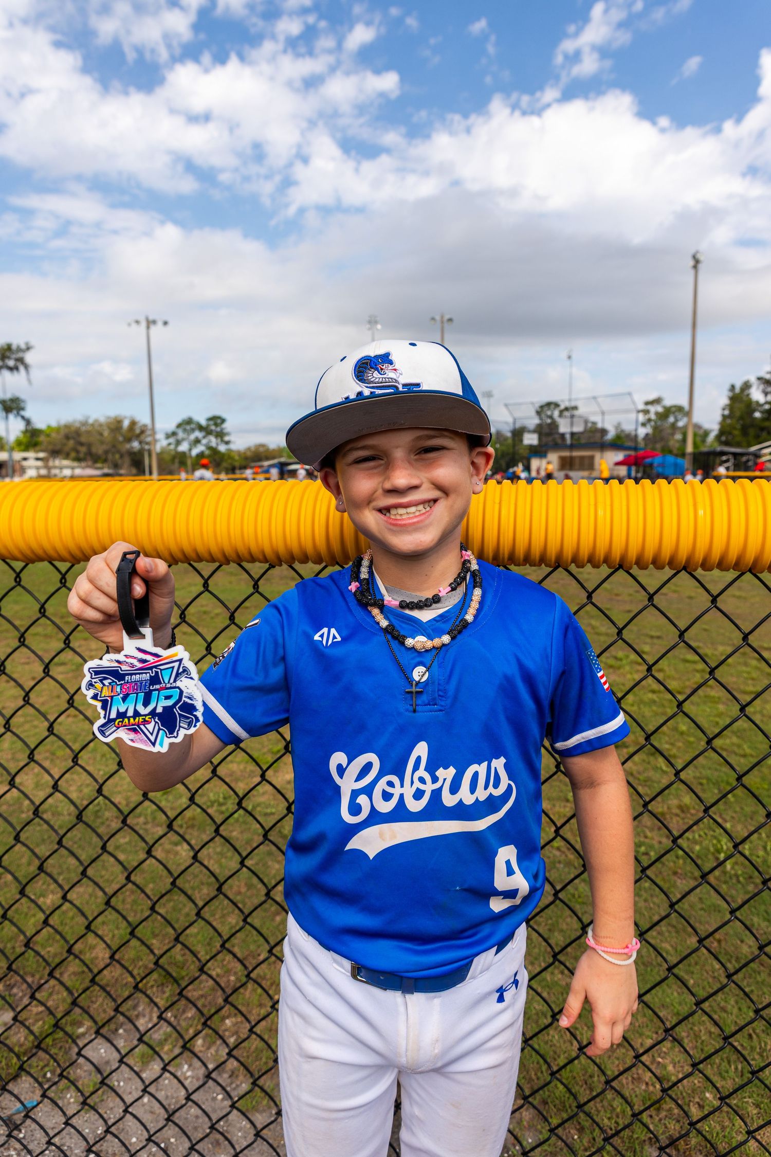 A young baseball player in a blue and white Coolers uniform stands by a chain link fence on a sunny day.