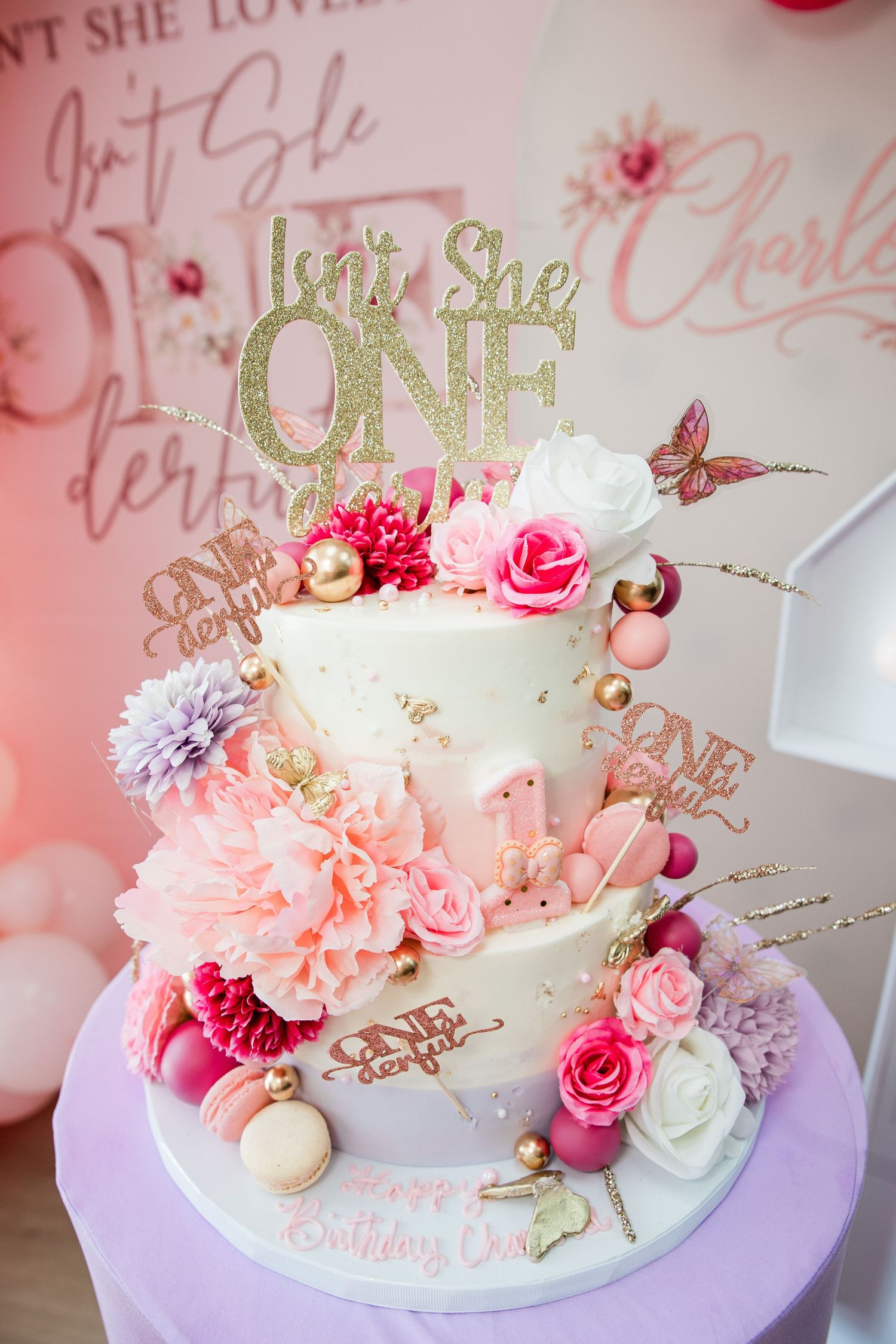 A two-tier white birthday cake with pink flowers, gold glitter topper reading Isn't She ONE, macarons, and butterflies for a first birthday.