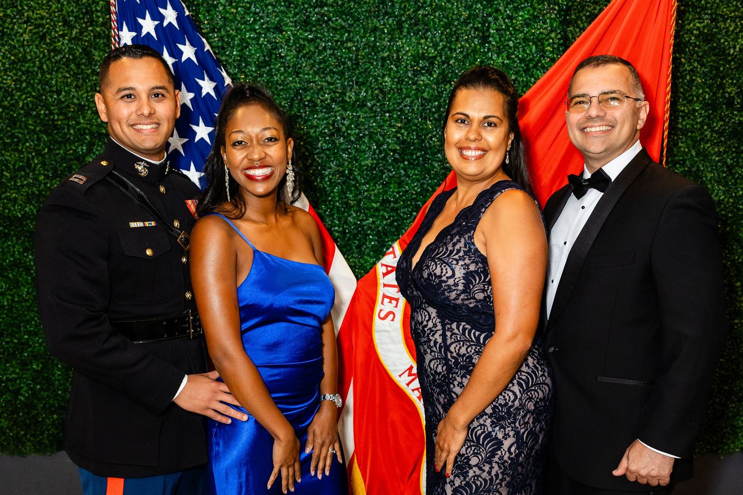 Four formally dressed individuals pose together in front of an American flag and green hedge wall at a military gala event.