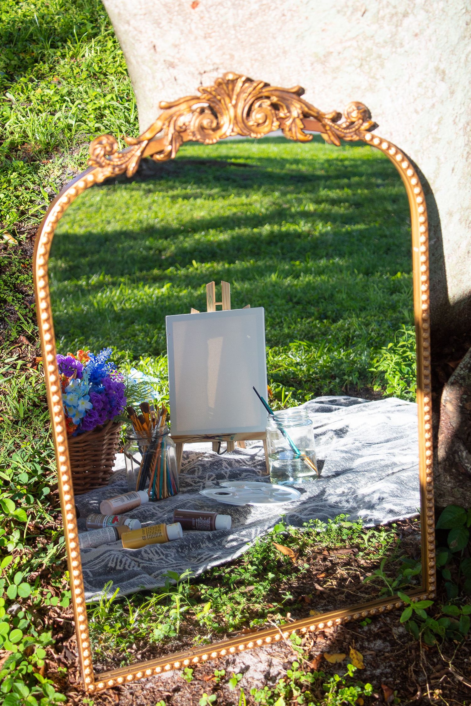 Ornate gold-framed mirror reflecting small easel setup on grass with flowers nearby.