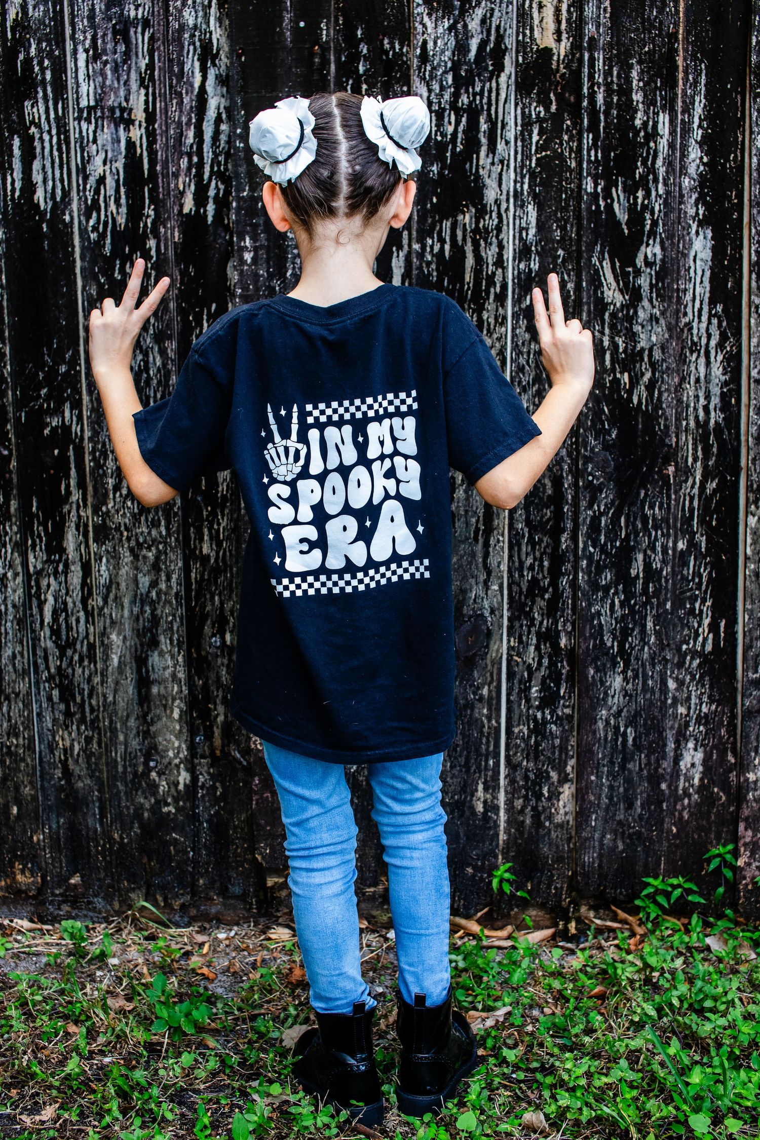 Child in spooky t-shirt and bows stands against dark wooden wall.