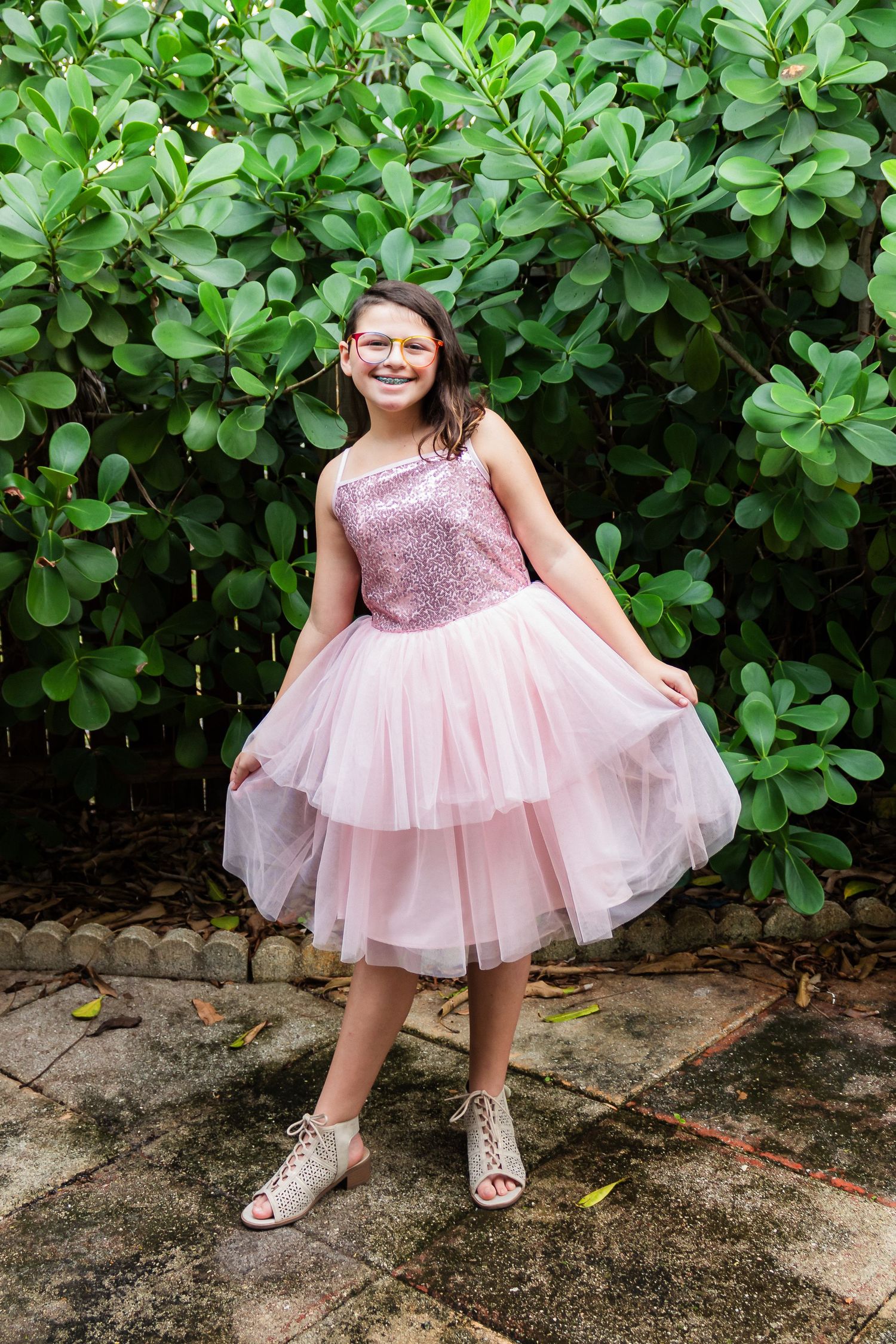 Young person in pink tulle dress poses in front of lush green foliage.