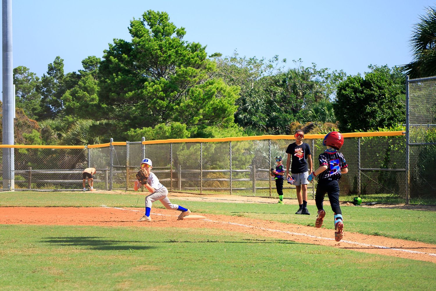 Baseball players compete in a youth league game on a well-maintained field with chain link fence backdrop.