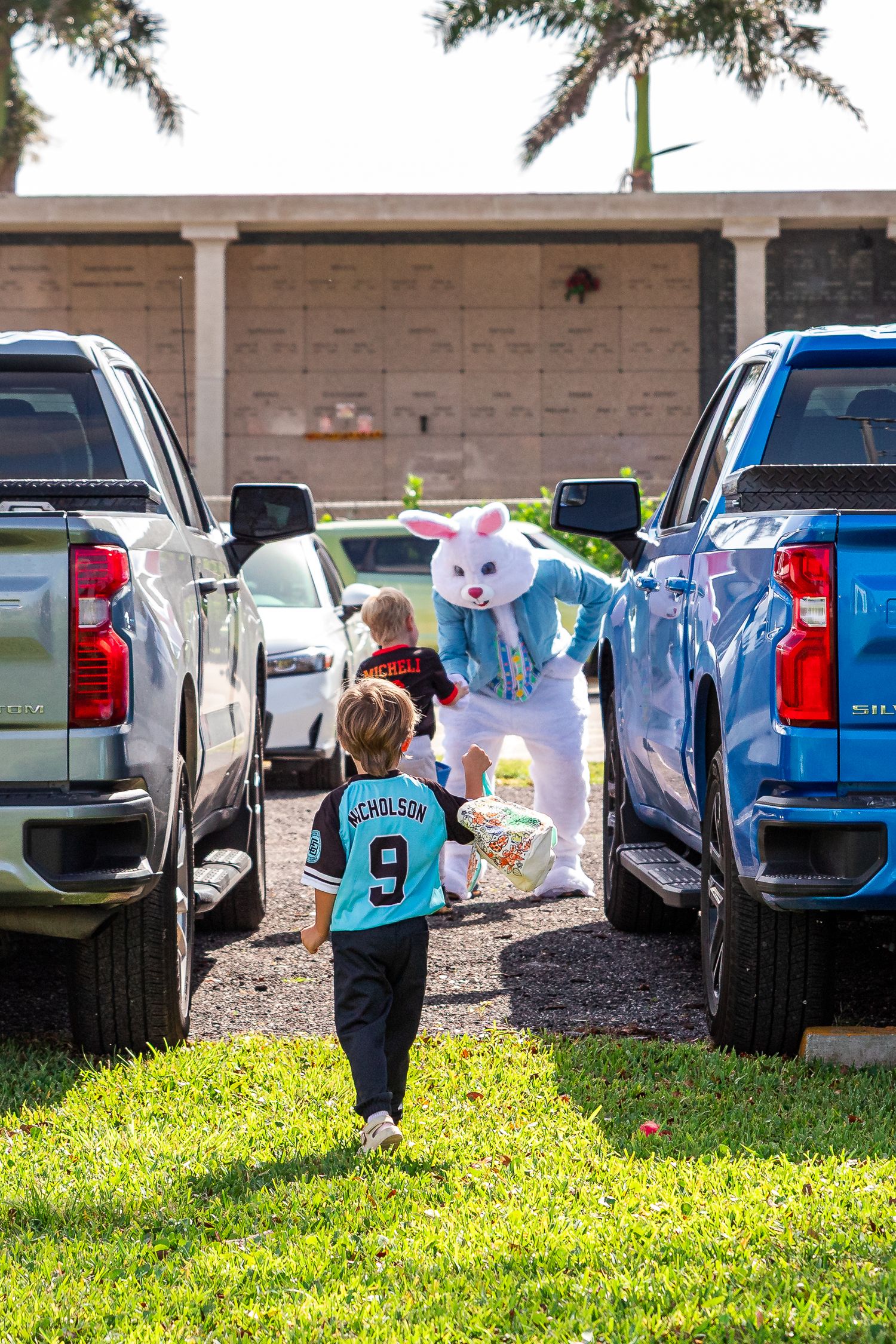A child in a baseball jersey approaches the Easter Bunny between parked trucks during an Easter egg hunt.