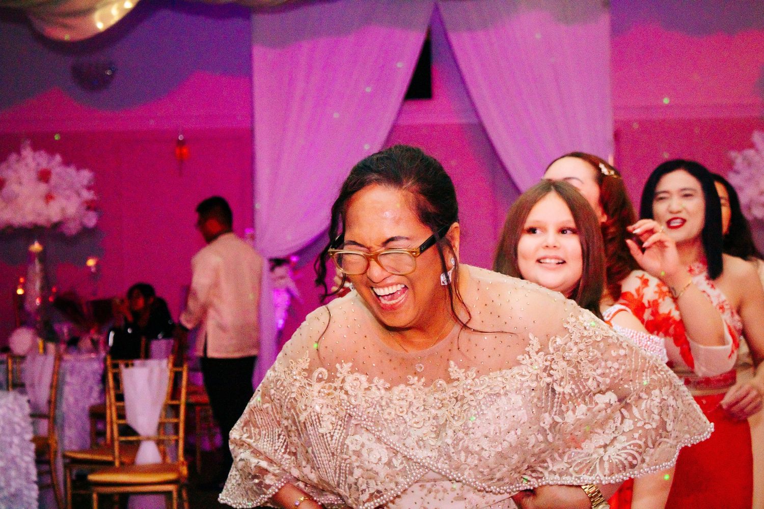 Wedding reception celebration with guests dancing under vibrant pink lighting and floral decor.