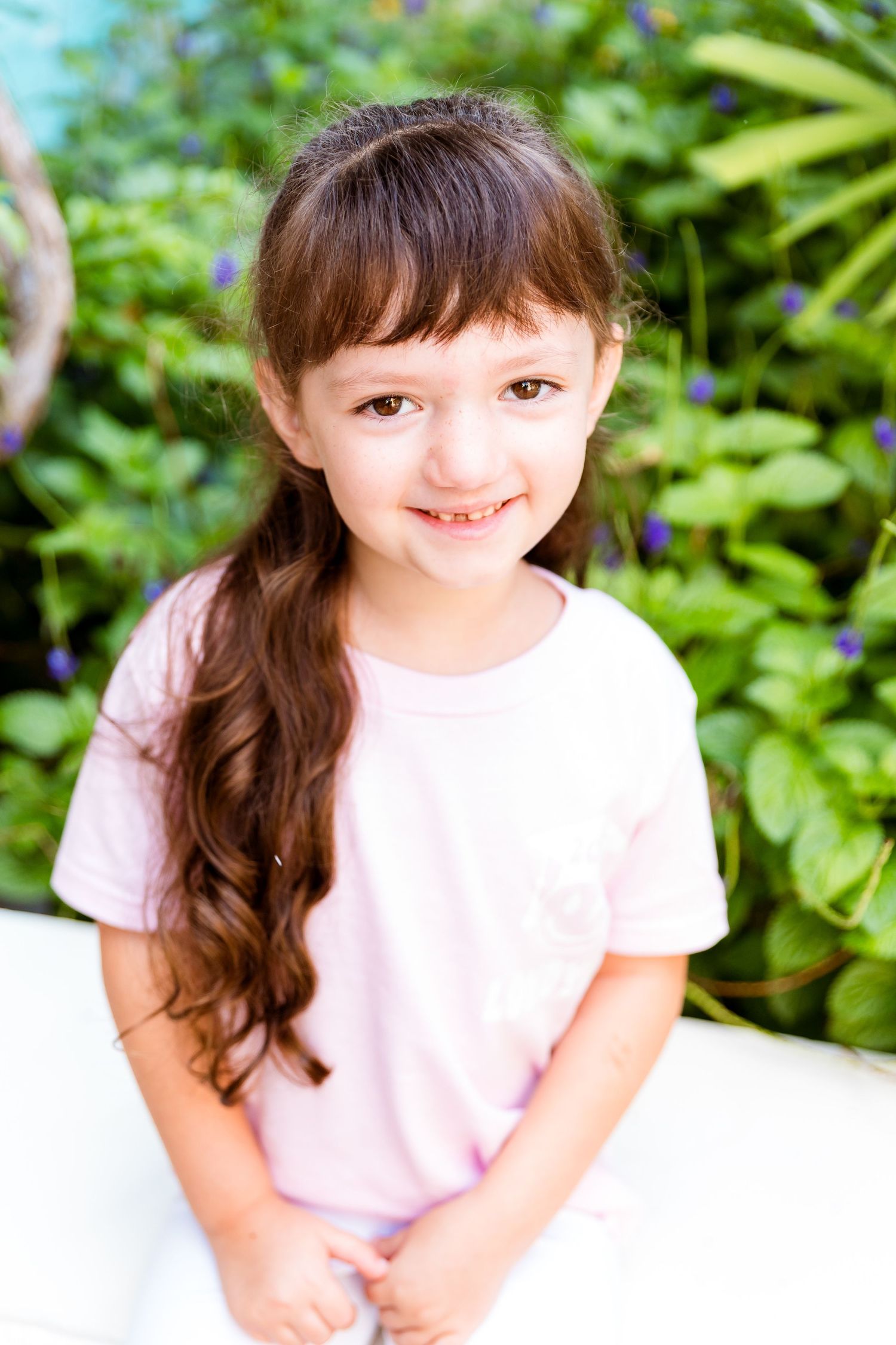 Child with long brown wavy hair wearing pink t-shirt sits outdoors against green foliage backdrop.