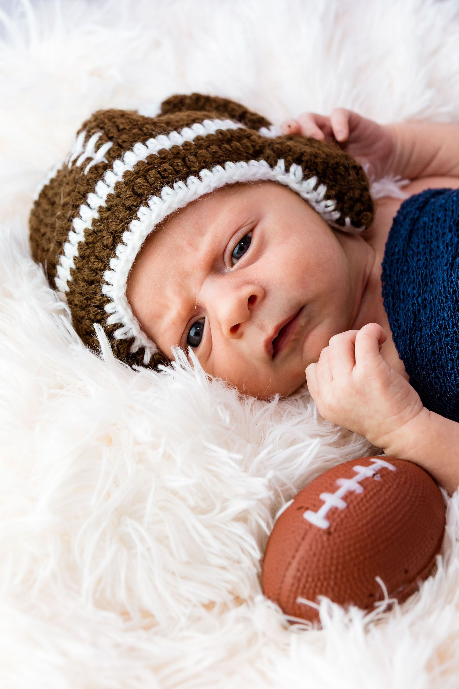 Newborn wearing football-themed knit hat lying on white fur with toy football.