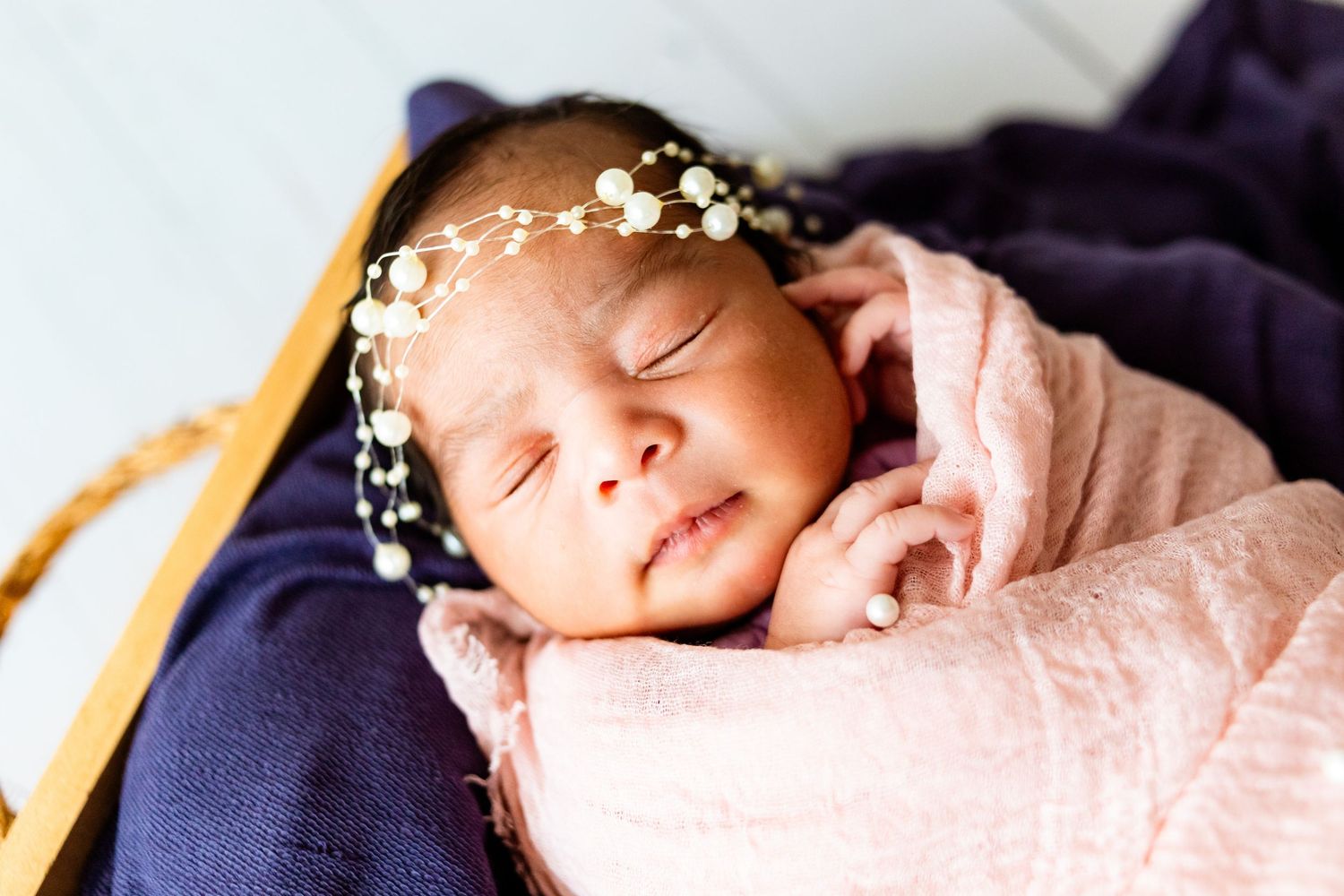 Sleeping newborn baby wrapped in pink swaddle with floral headband in a wooden basket.