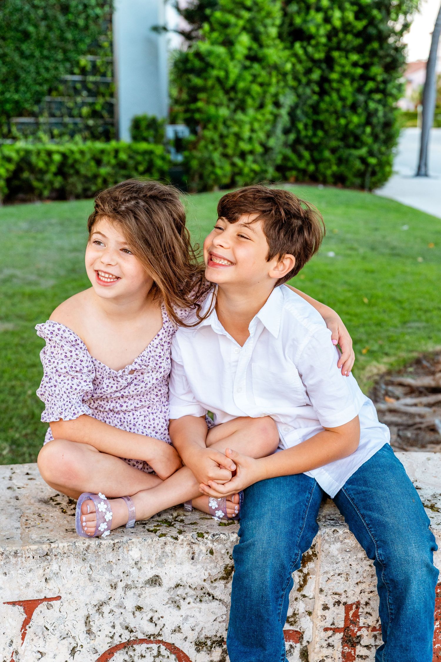 Two happy children sitting outdoors on a stone ledge, smiling and hugging in front of lush green hedges.