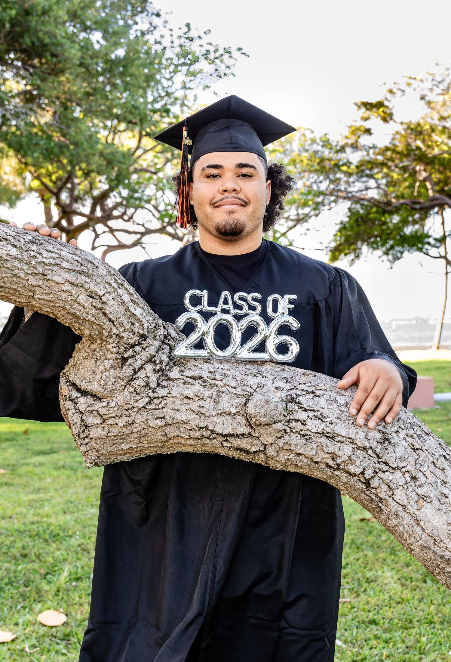 A young man in black graduation cap and gown poses by a tree, wearing a Class of 2026 shirt outdoors.