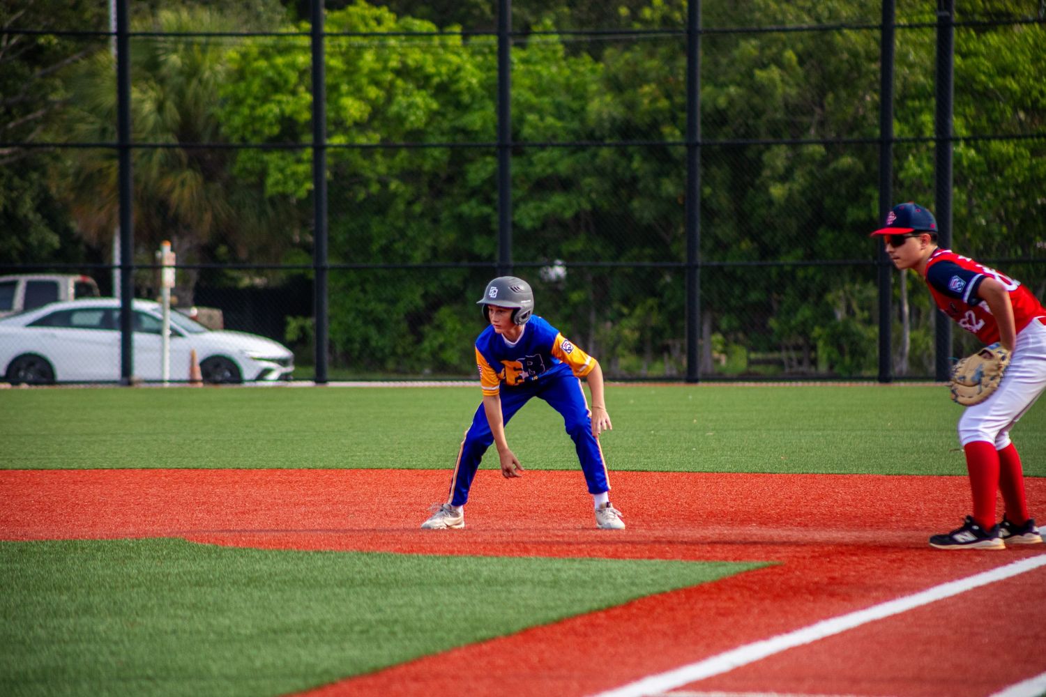 Young baseball player in blue and orange uniform leads off base during a youth baseball game on a red turf field.