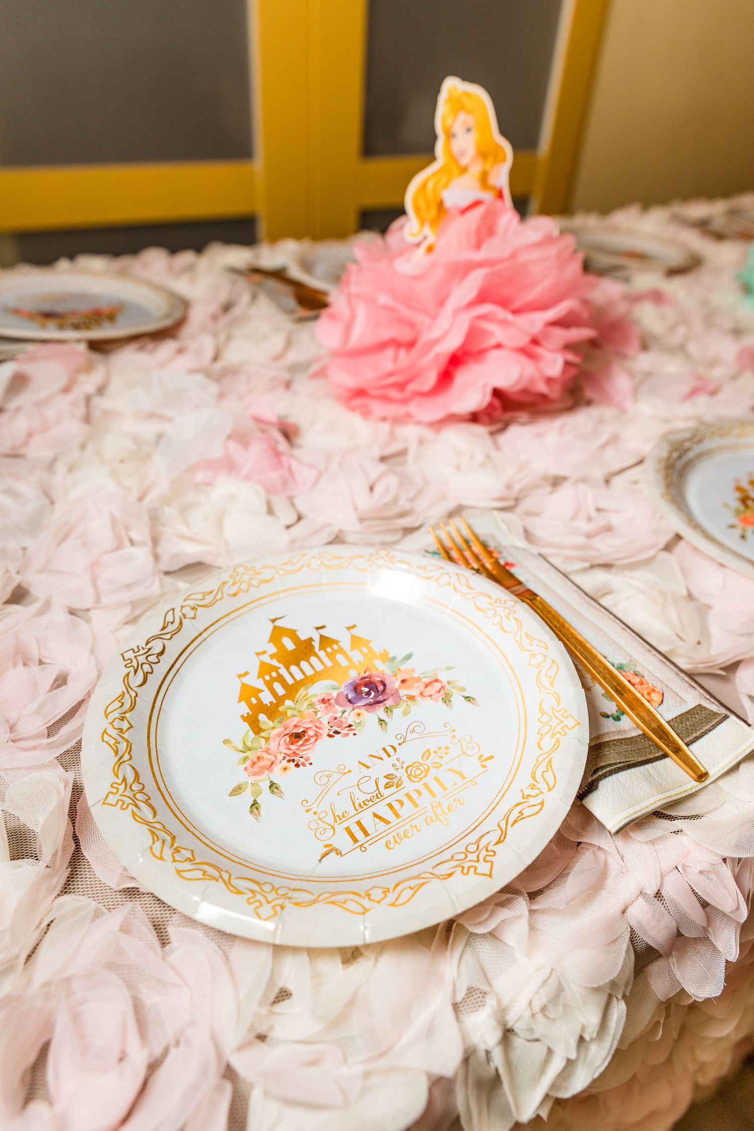 Pink and gold princess-themed table setting with delicate floral plates and ruffled tablecloth at a special event.