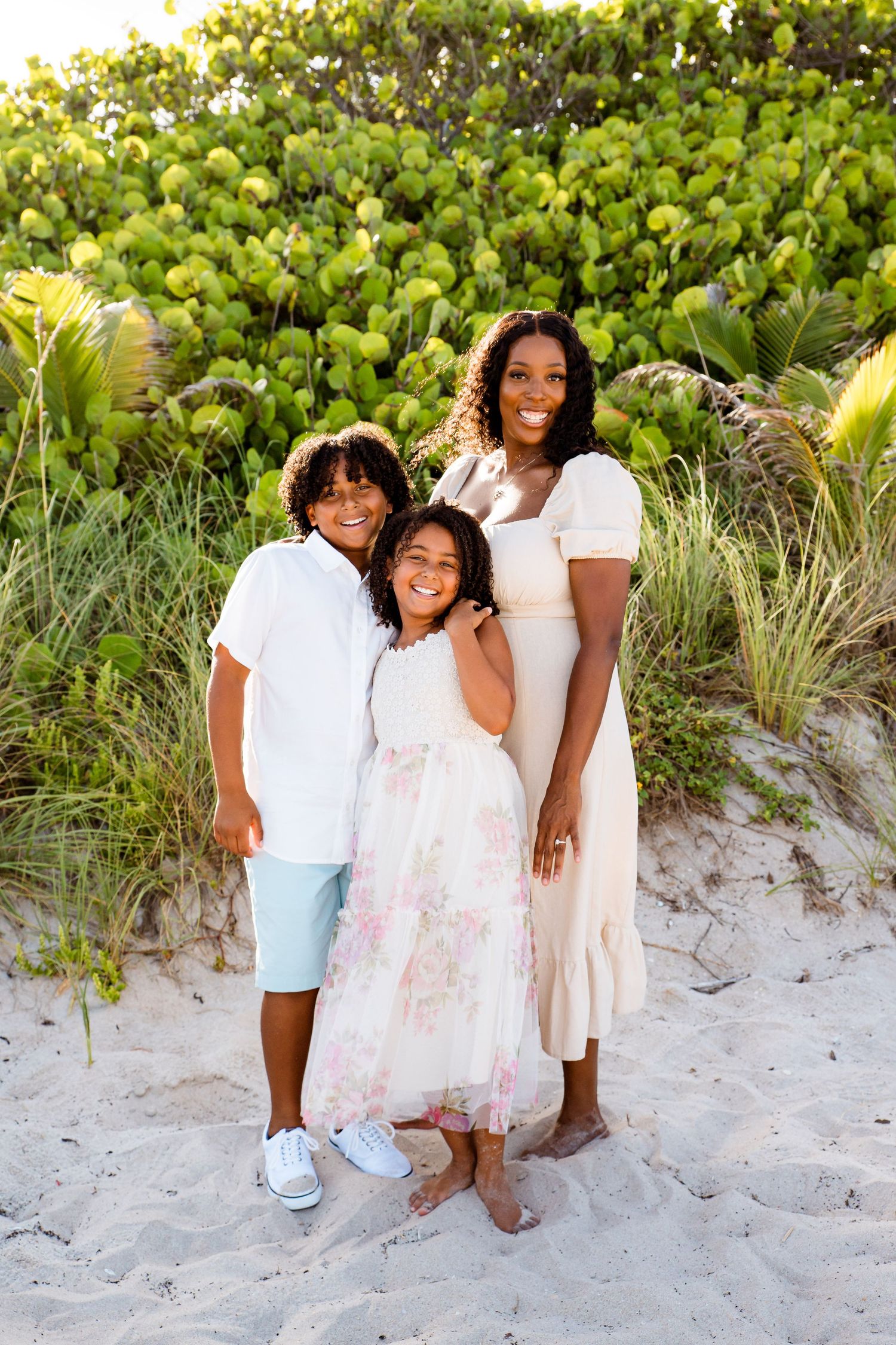 Three people pose together on a sandy beach with lush green sea grape plants in the background.