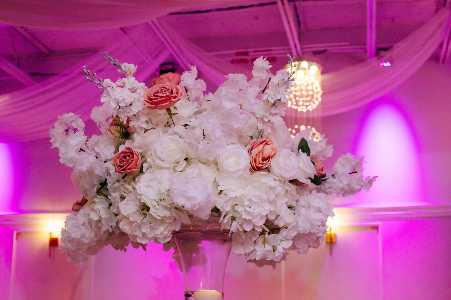 Luxurious white floral arrangements with crystal chandeliers under pink and purple event lighting.