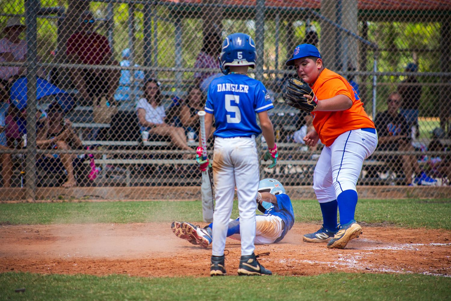 Baseball players in blue and orange uniforms interact at first base during a game on a dirt diamond field.