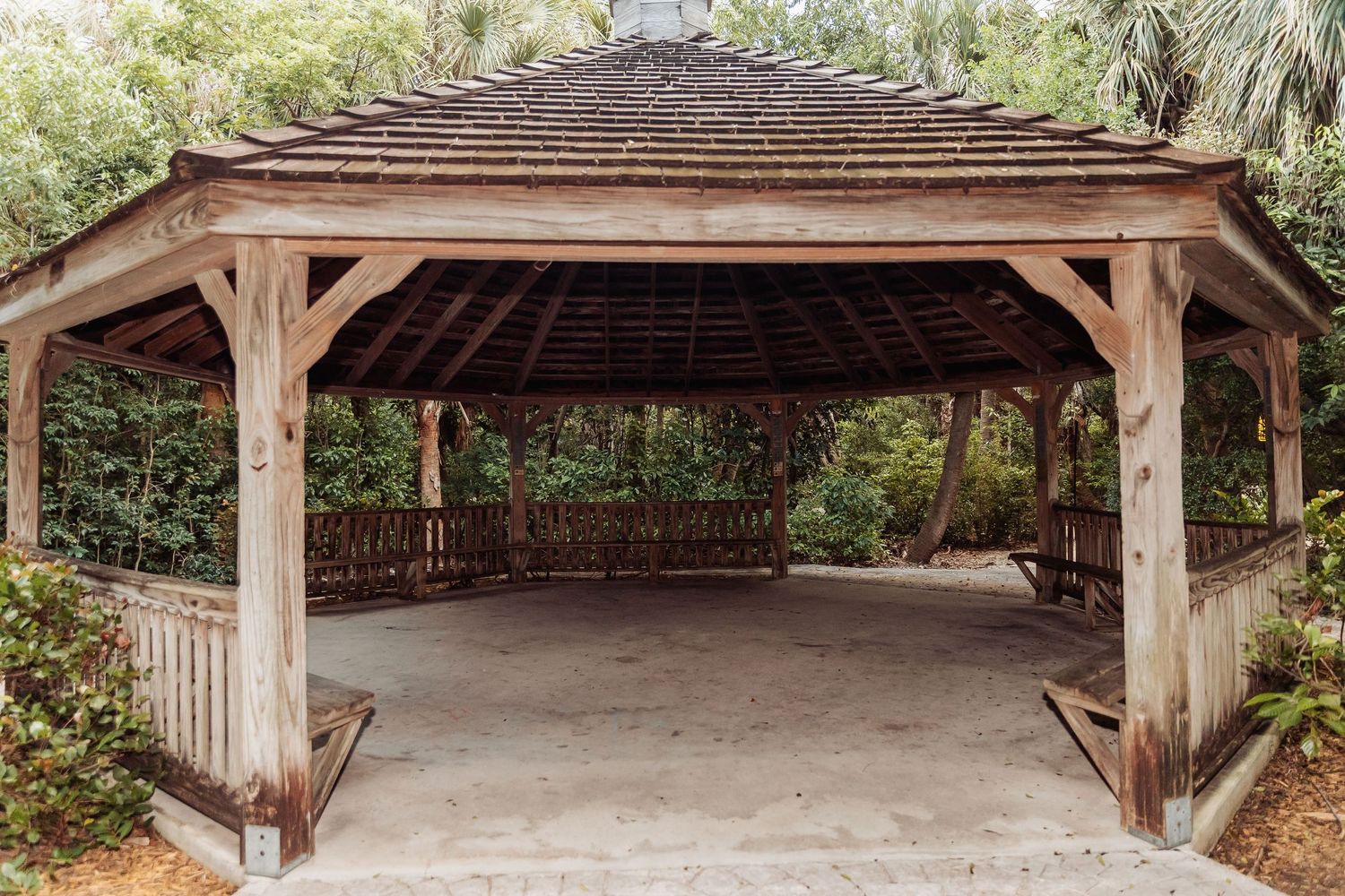 A rustic wooden pavilion with a brown shingled roof stands in a lush garden setting.
