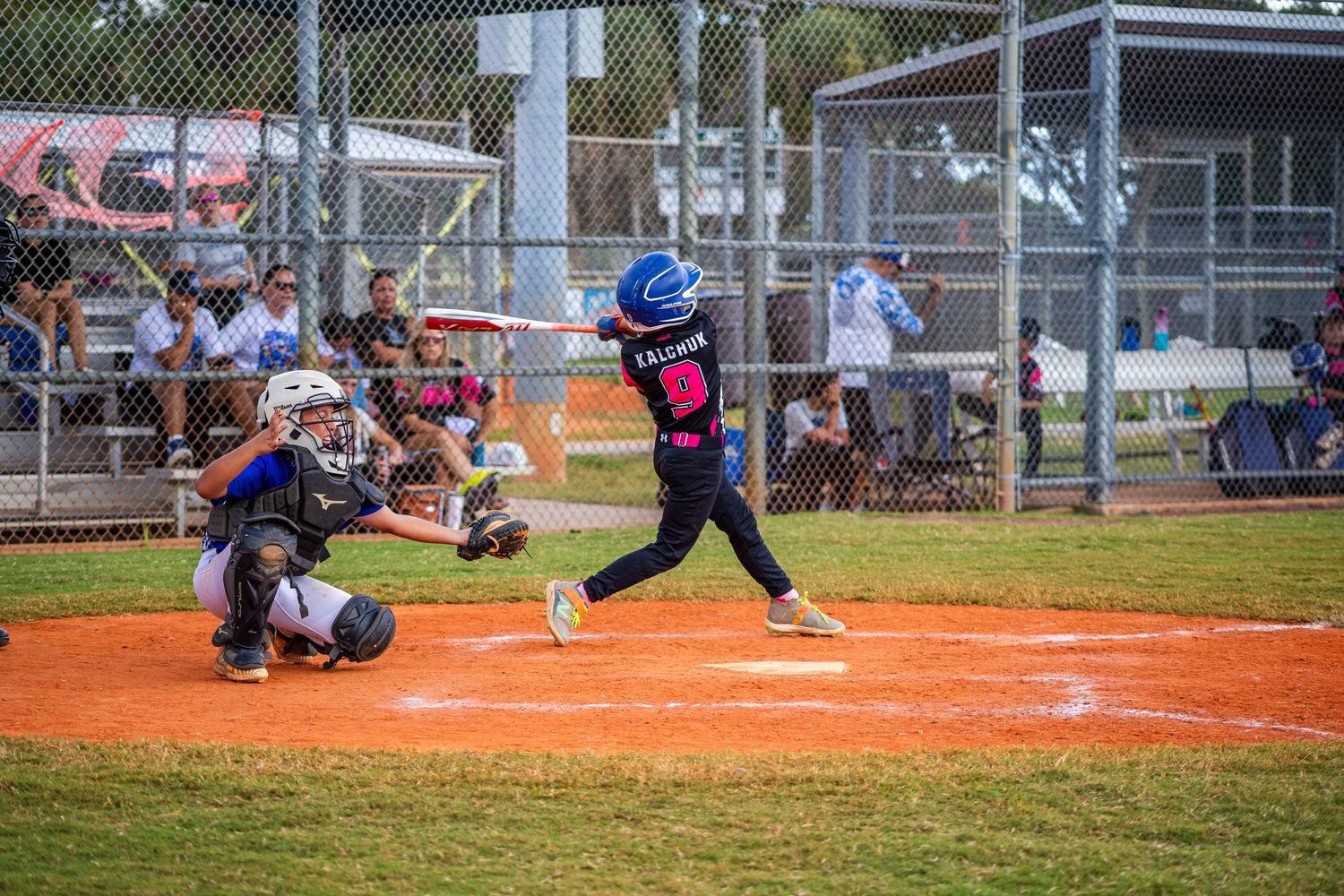 Baseball player in blue uniform swings bat at home plate during youth league game on dirt field.