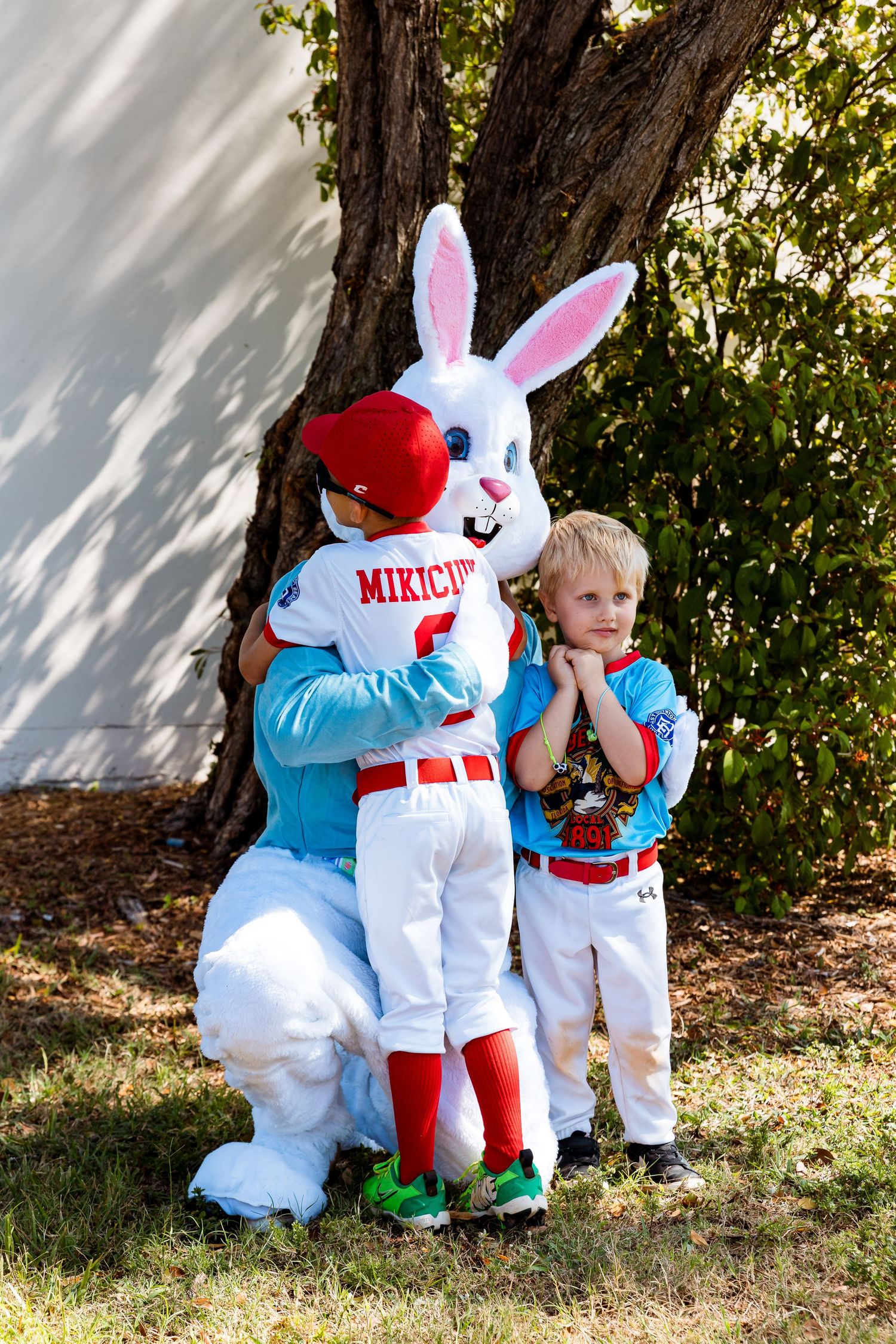 Someone in an Easter Bunny costume poses with a young child in matching baseball uniforms outdoors by a tree.