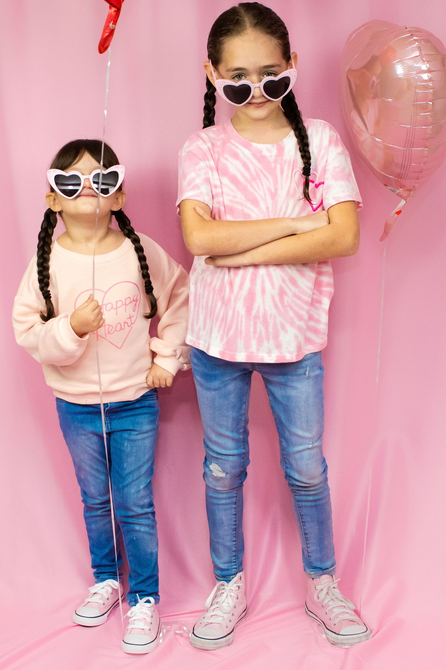 Two children wearing heart sunglasses and matching pink outfits pose against pink backdrop.