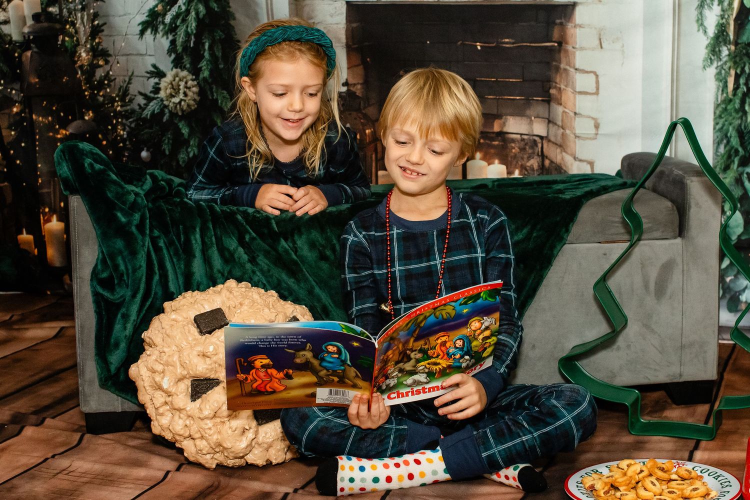 Two children in matching navy pajamas share a storybook while sitting on a green couch.