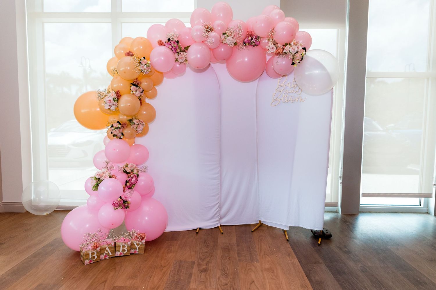 Pink and peach balloon arch decoration with floral accents against a white backdrop and hardwood floor.