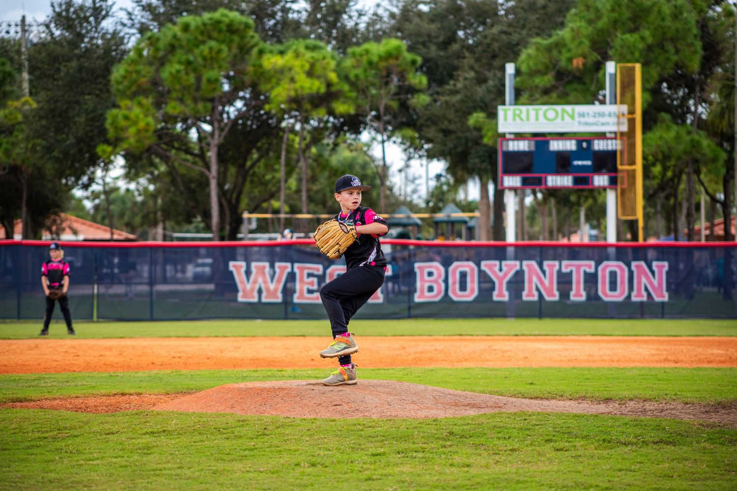 Baseball action shot on field with scoreboard and trees visible in background.
