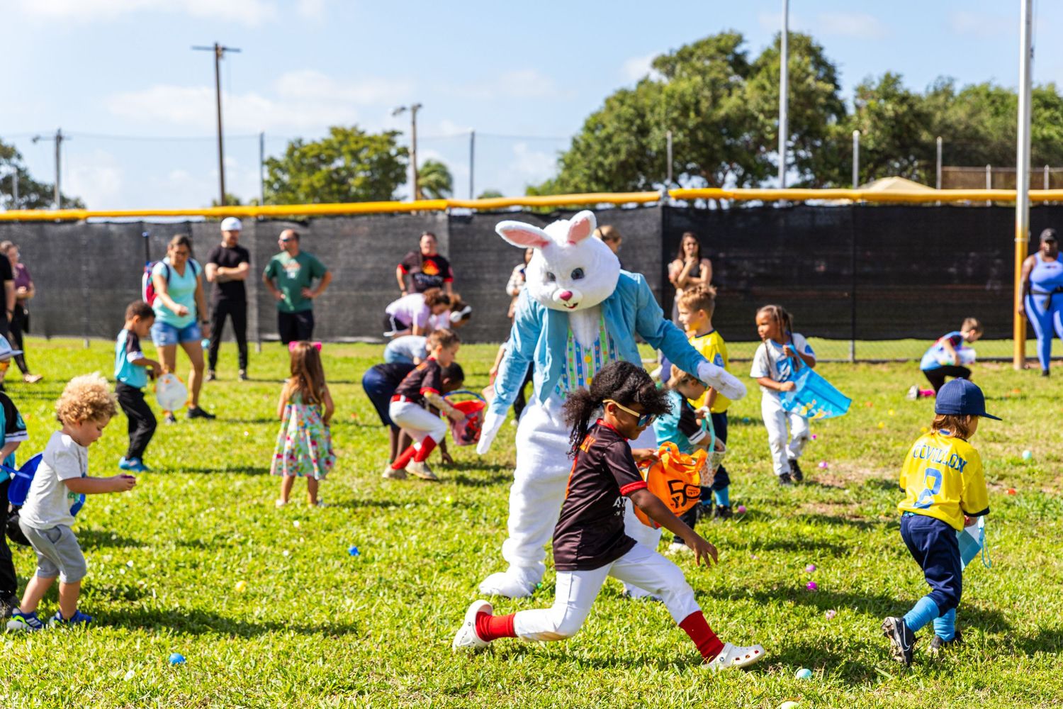 Easter Bunny costume character interacts with children during an outdoor Easter egg hunt on a sunny green field.