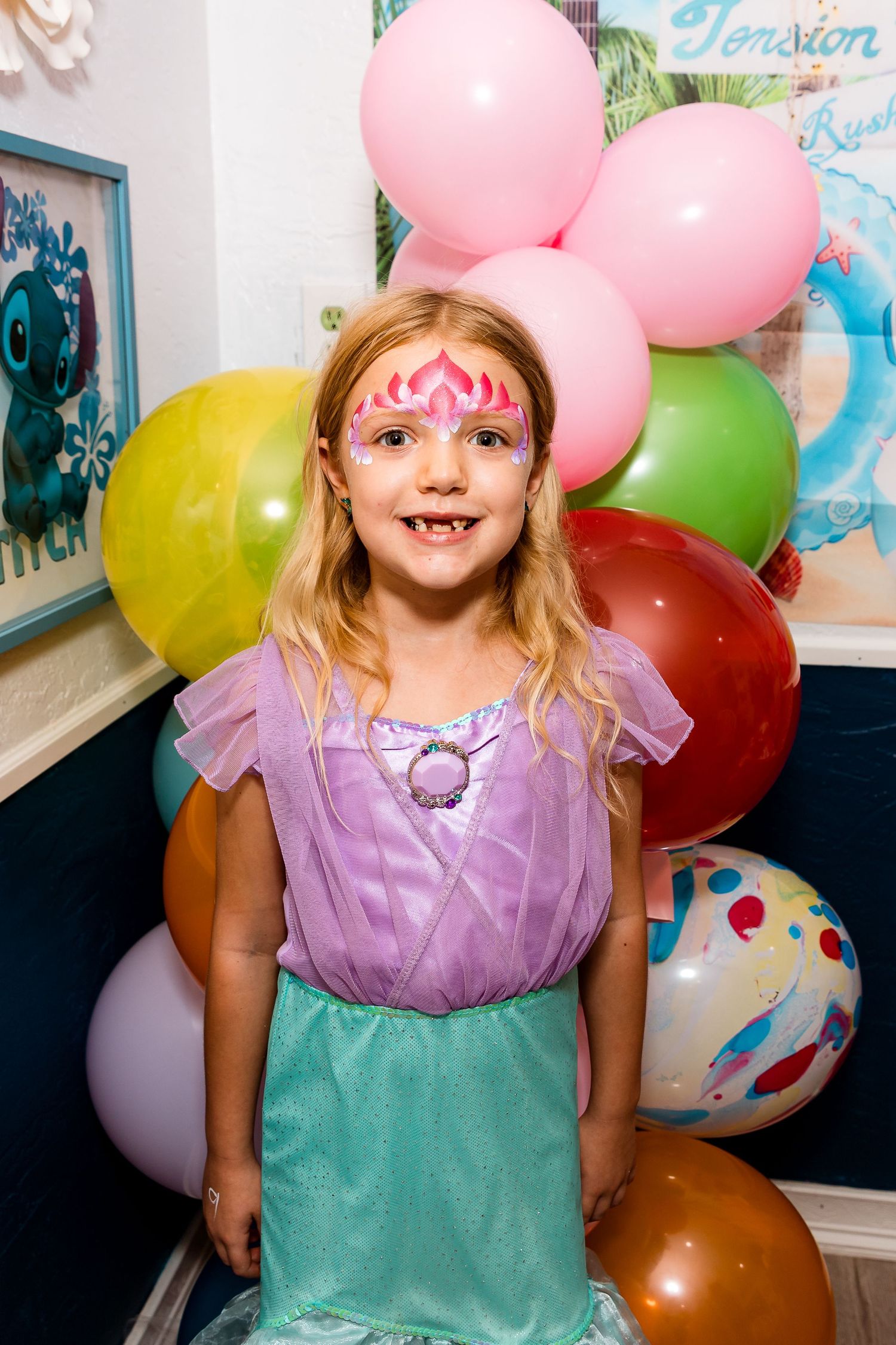 A child in a purple and teal costume dress poses with face paint in front of colorful balloons.