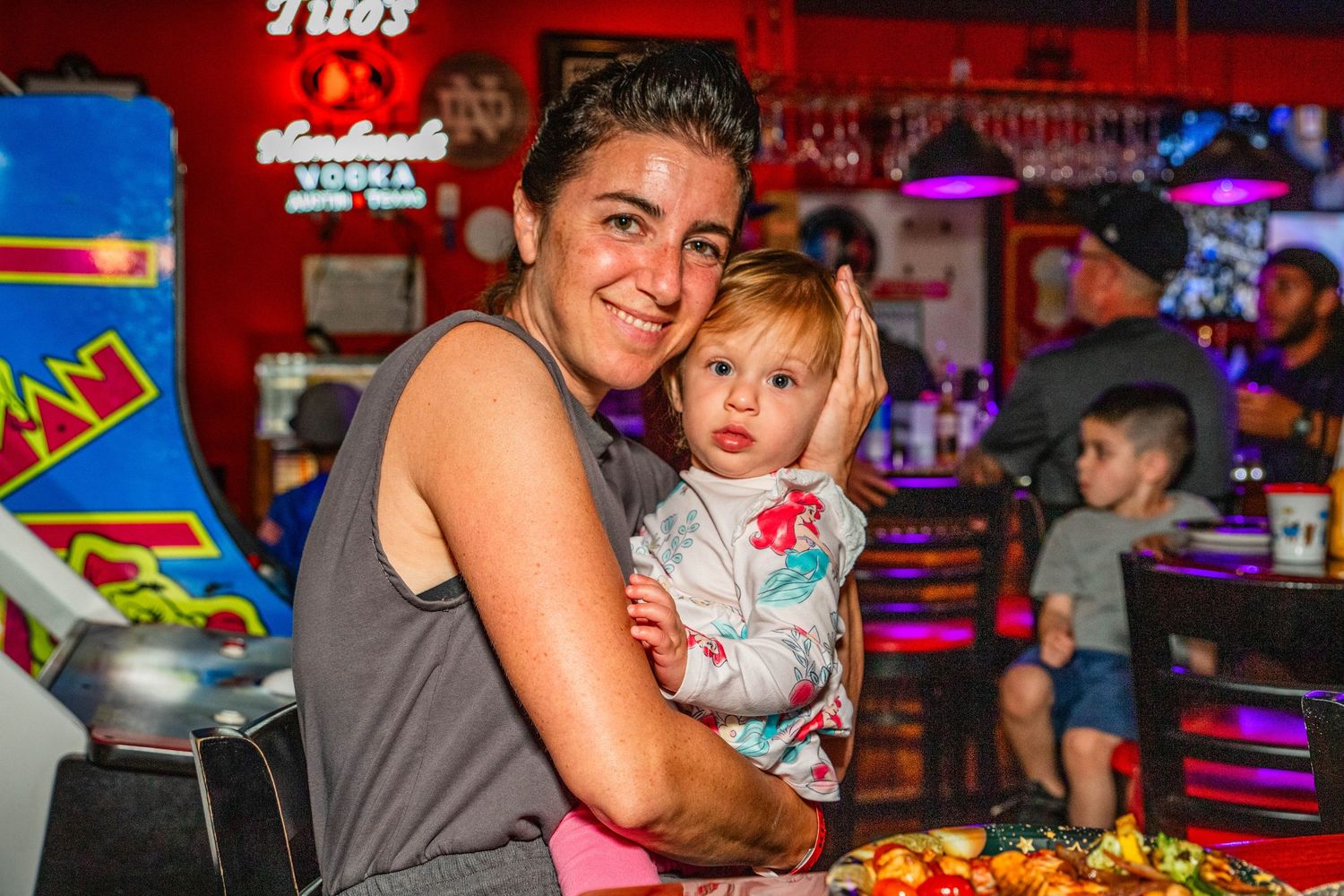 A woman holds a baby at a colorful bar with arcade games and neon signs in the background.