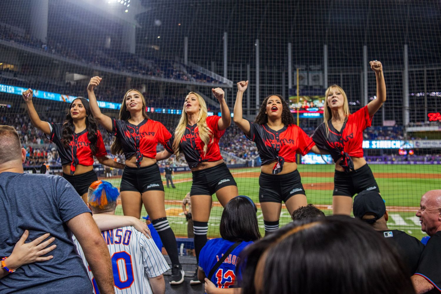Cheerleaders in red tops perform for crowd at baseball stadium during night game.