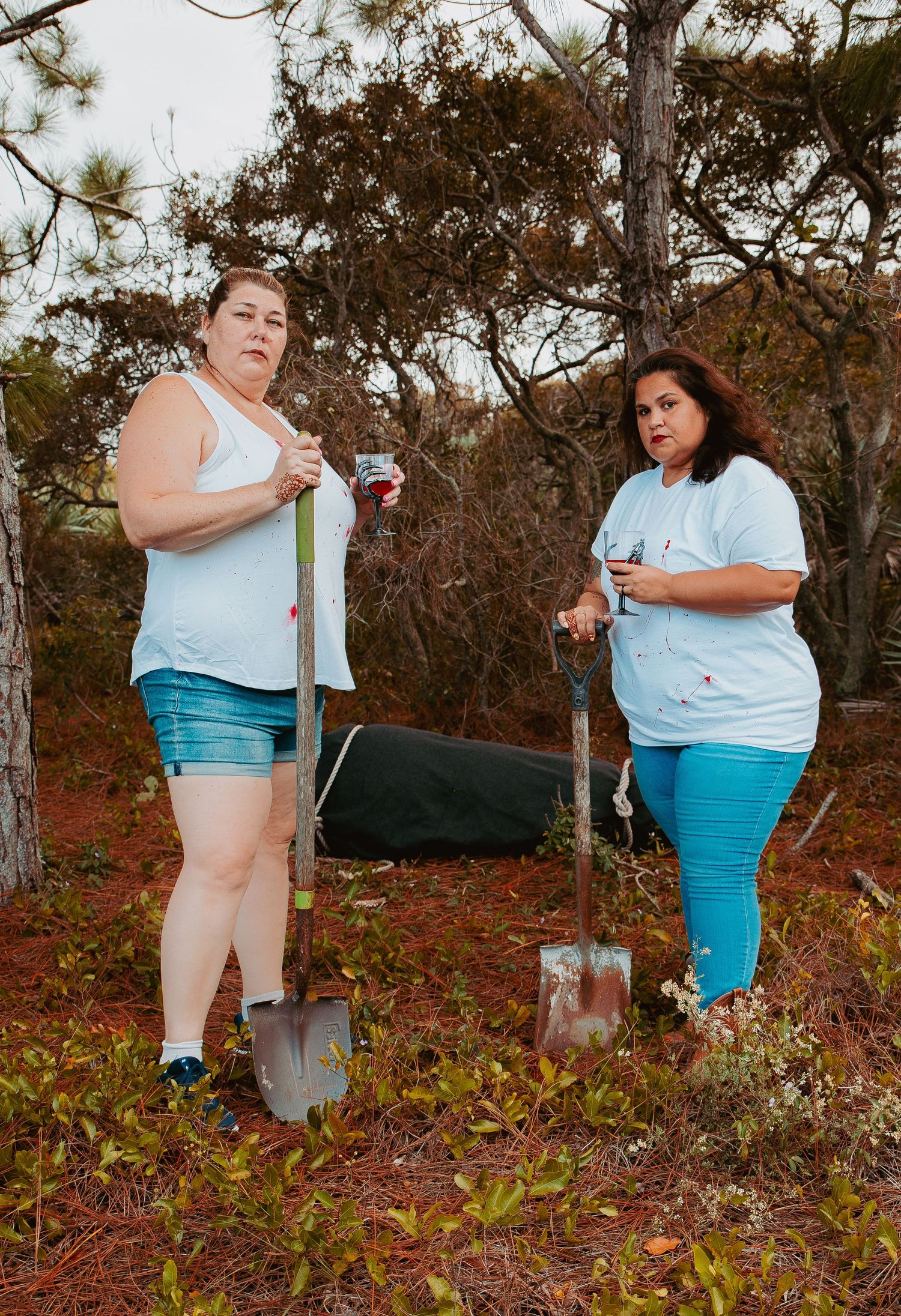 Two people stand in a wooded area holding shovels and water bottles during what appears to be a gardening activity.