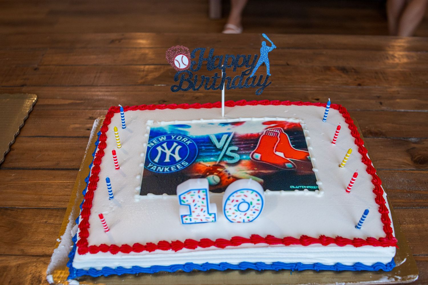 Long rectangular celebratory cake decorated in red, white and blue colors displayed on wooden table with sports theme.