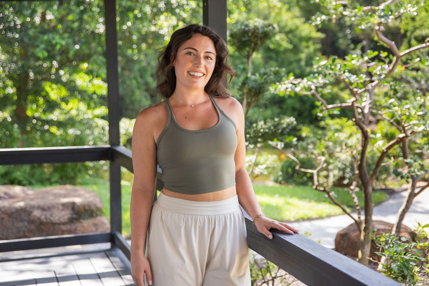 A woman in athletic wear stands confidently on an outdoor deck surrounded by lush greenery and trees.
