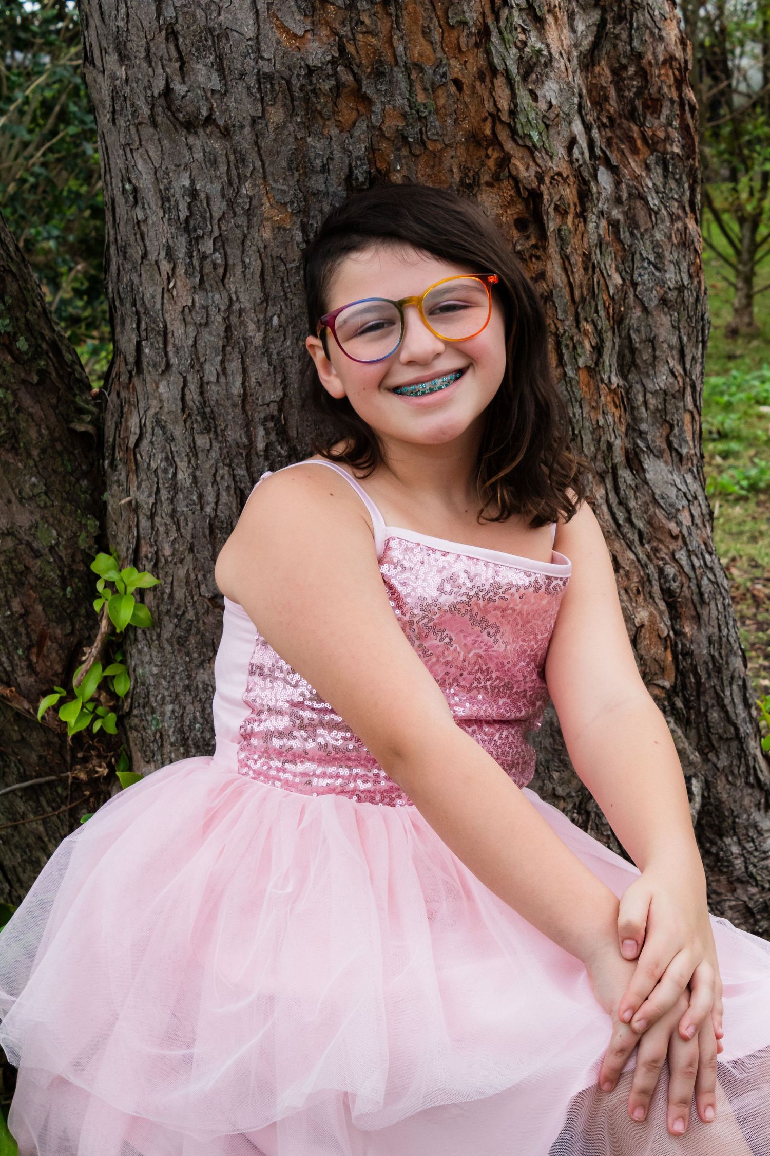 A young person in a pink sequined dress with tulle skirt and glasses poses against a tree trunk outdoors.