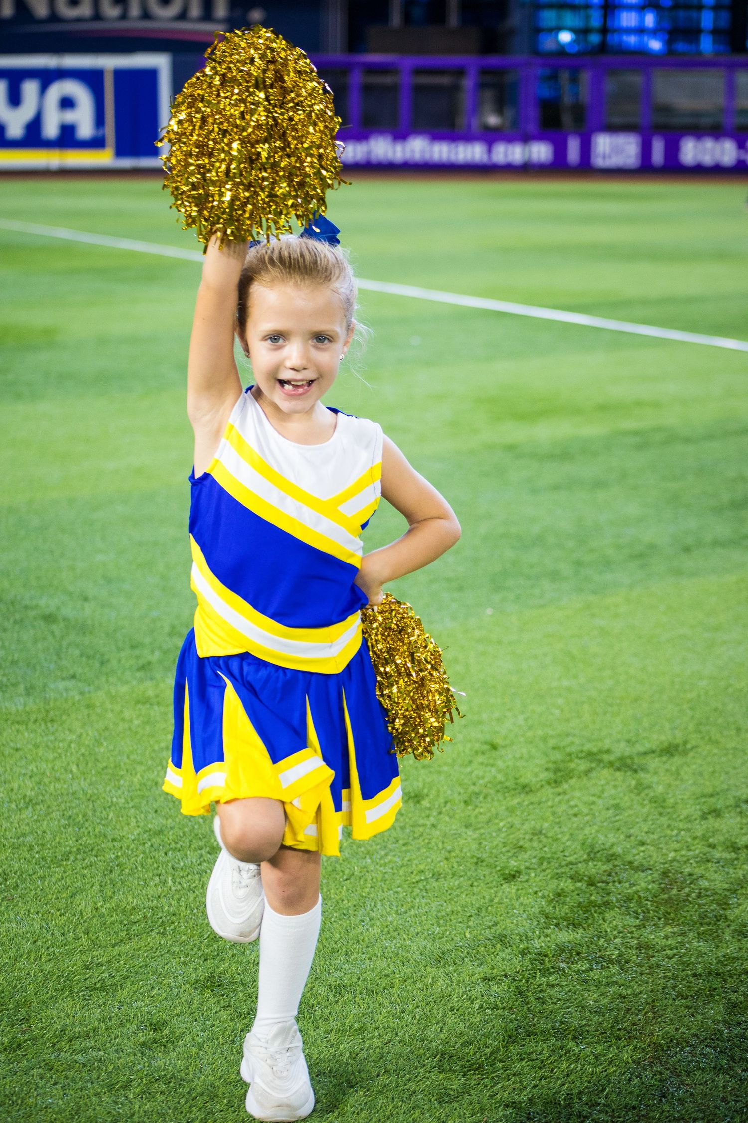 Cheerleader in blue and yellow uniform holds gold pom poms on sports field.