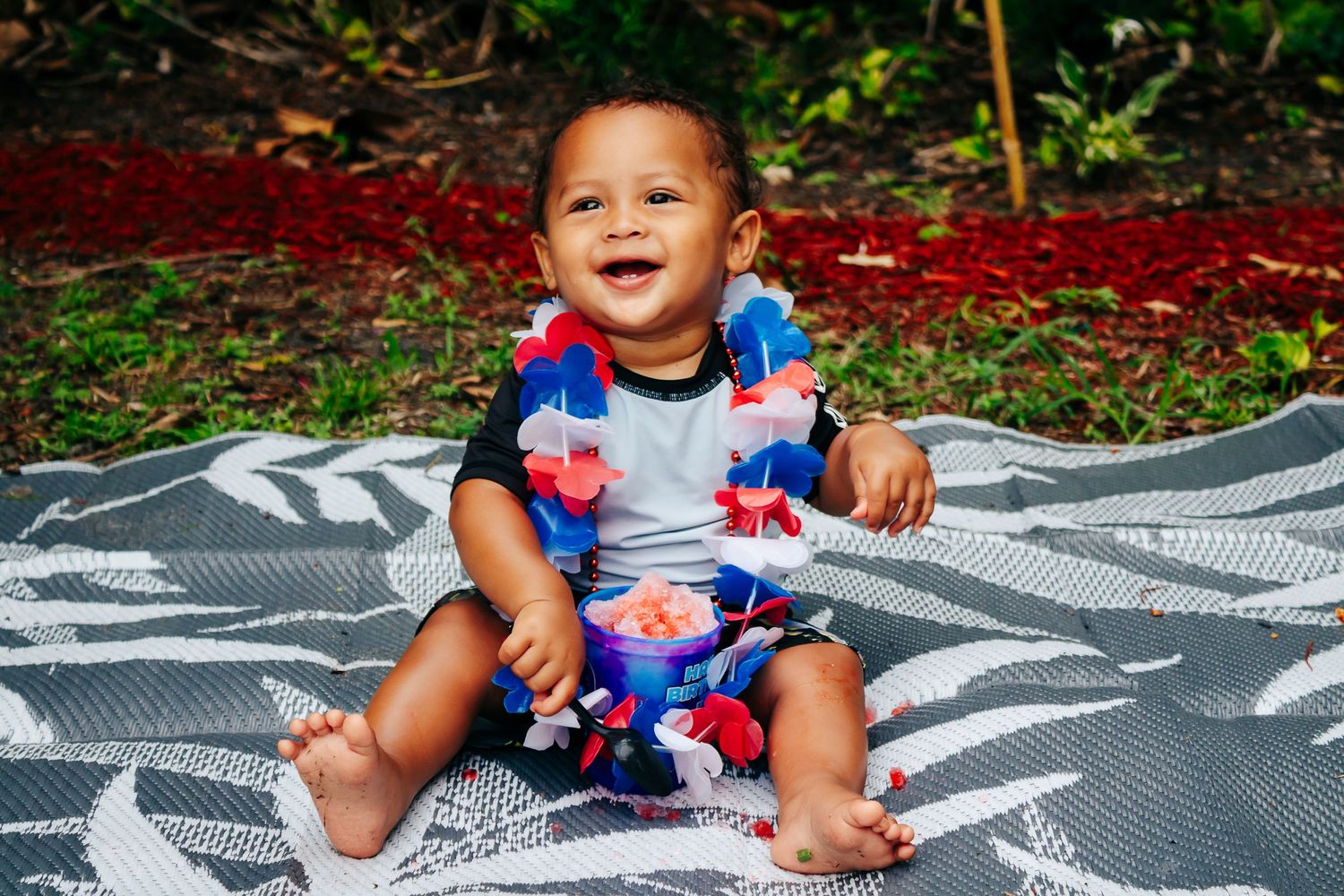 Baby wearing a floral outfit sitting on a patterned blanket outdoors.