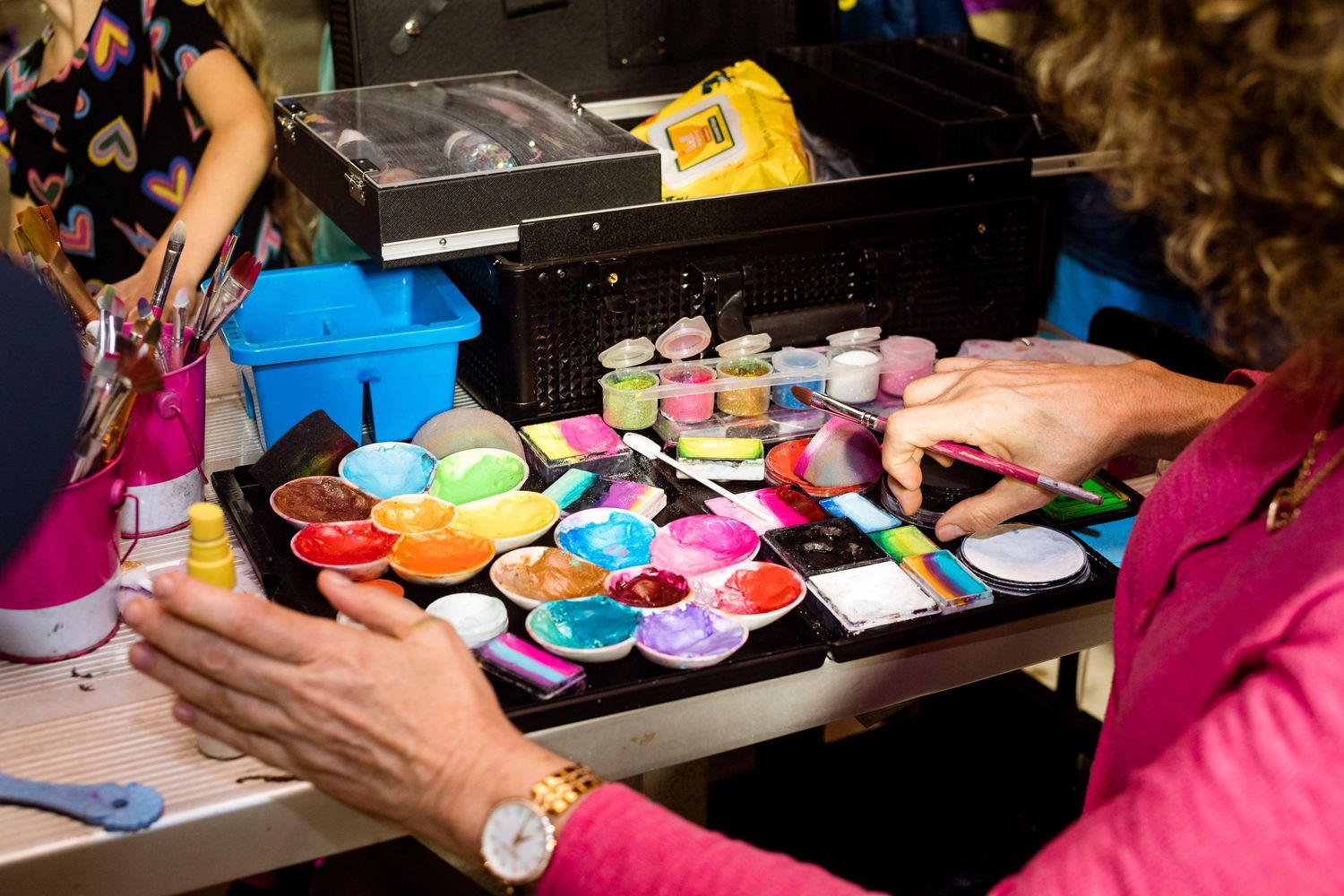 Hands arrange colorful face painting supplies and palettes on a black table.
