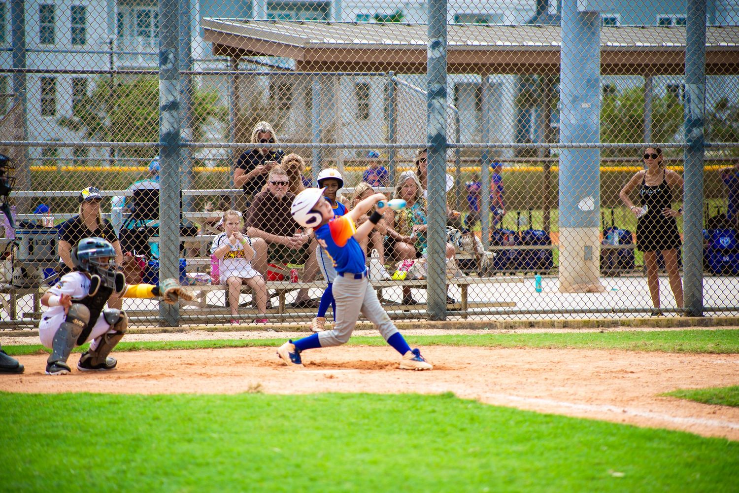 A baseball batter swings at a pitch during a youth baseball game with spectators watching.