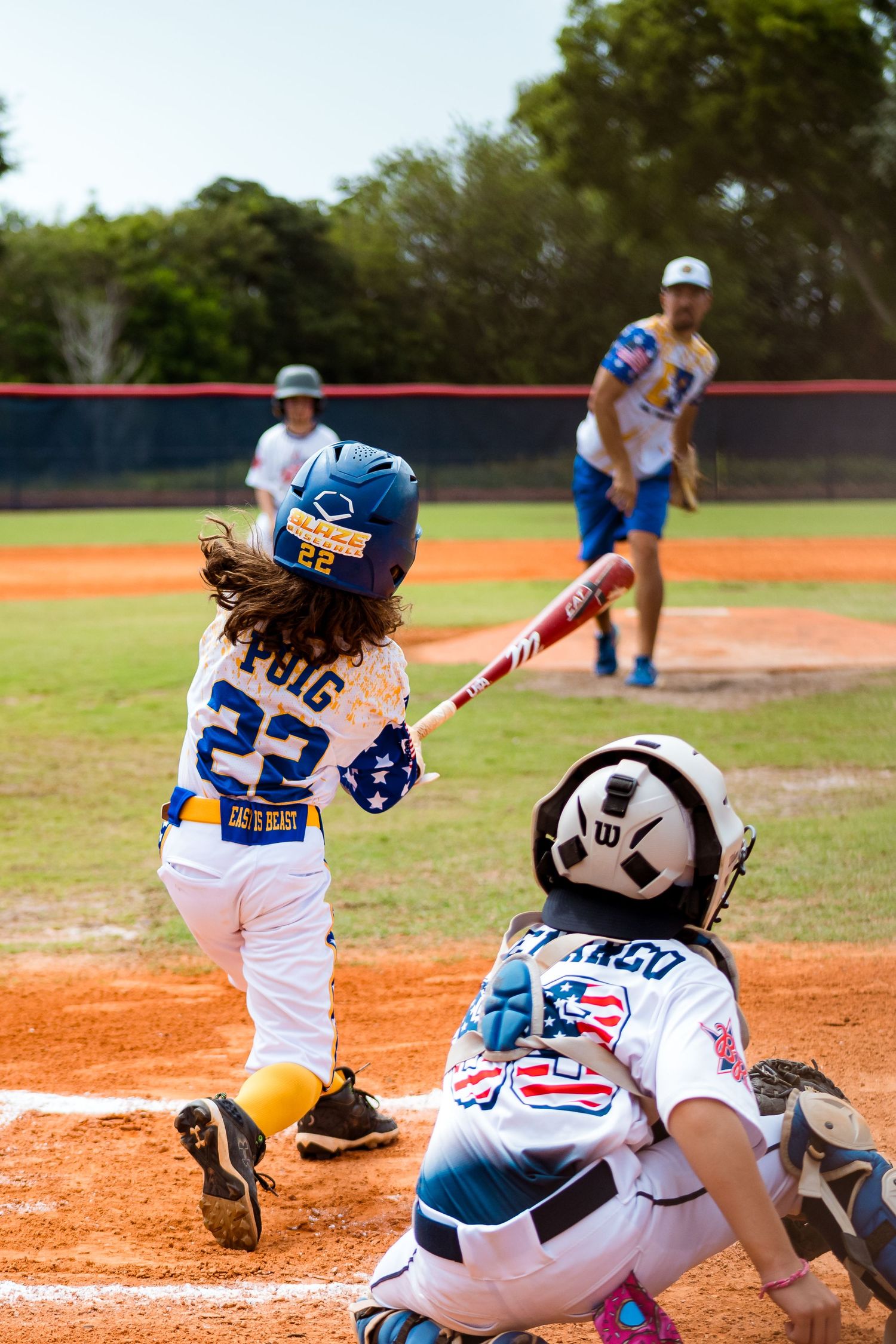 Youth baseball player swings a red bat at home plate during a little league game while the catcher crouches behind.