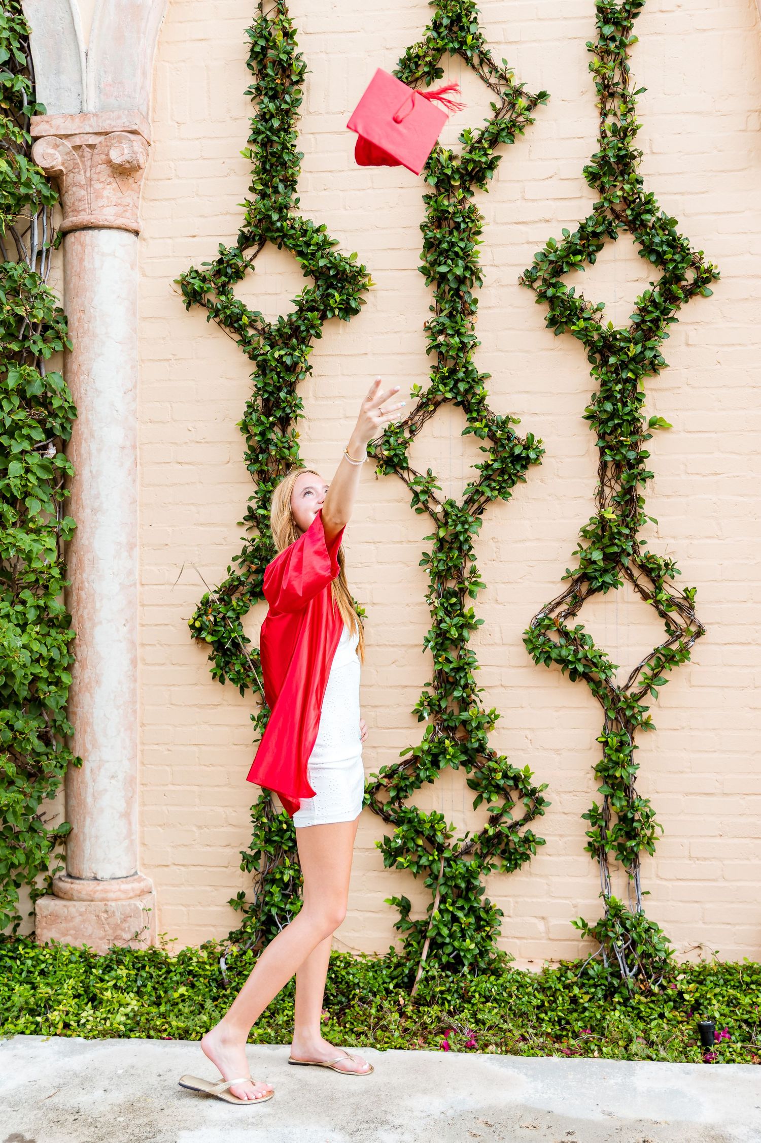 Graduate in white dress tossing red graduation cap against decorative ivy wall pattern.