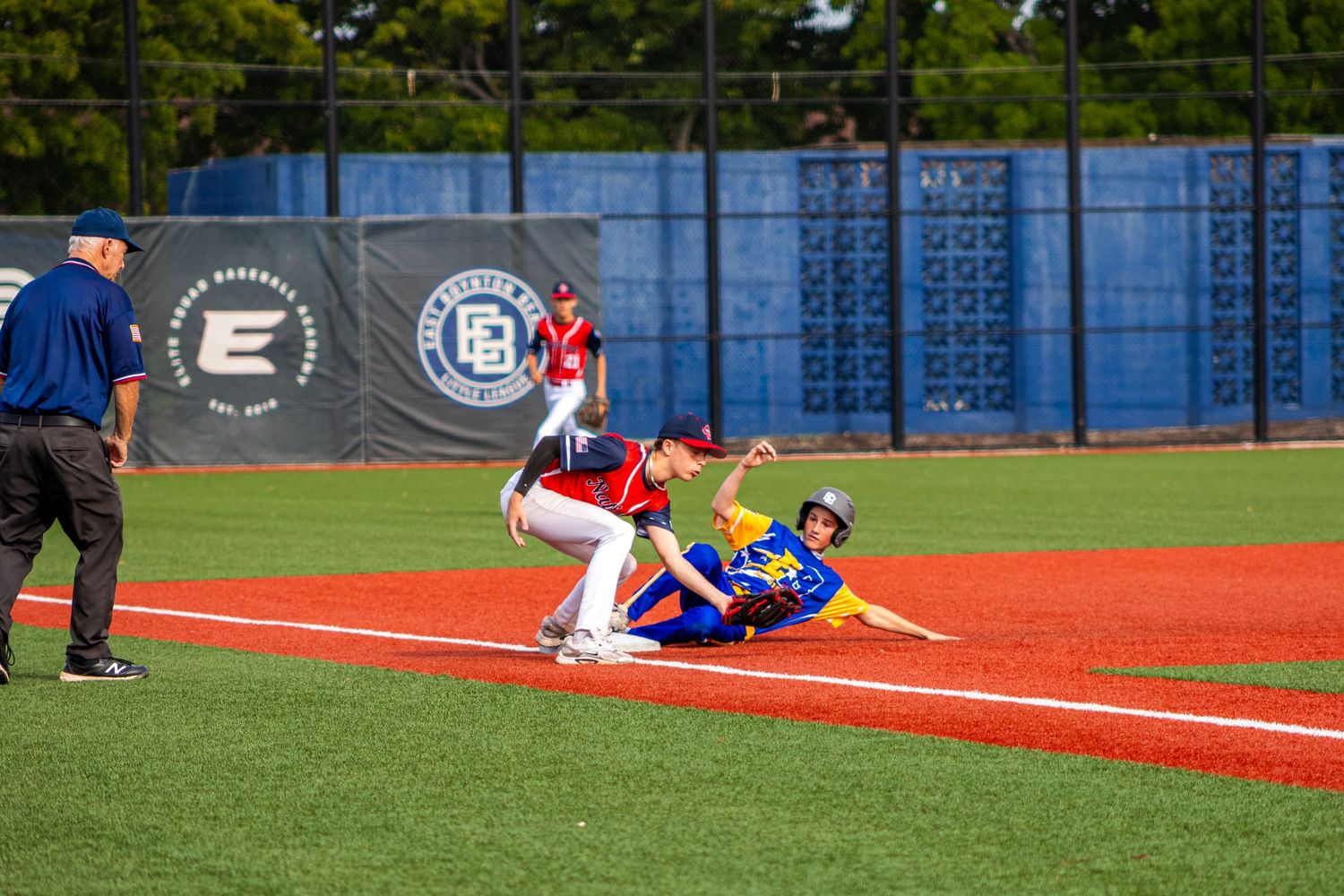 Baseball player sliding into third base while fielder attempts tag during a sunny outdoor game on red clay field.