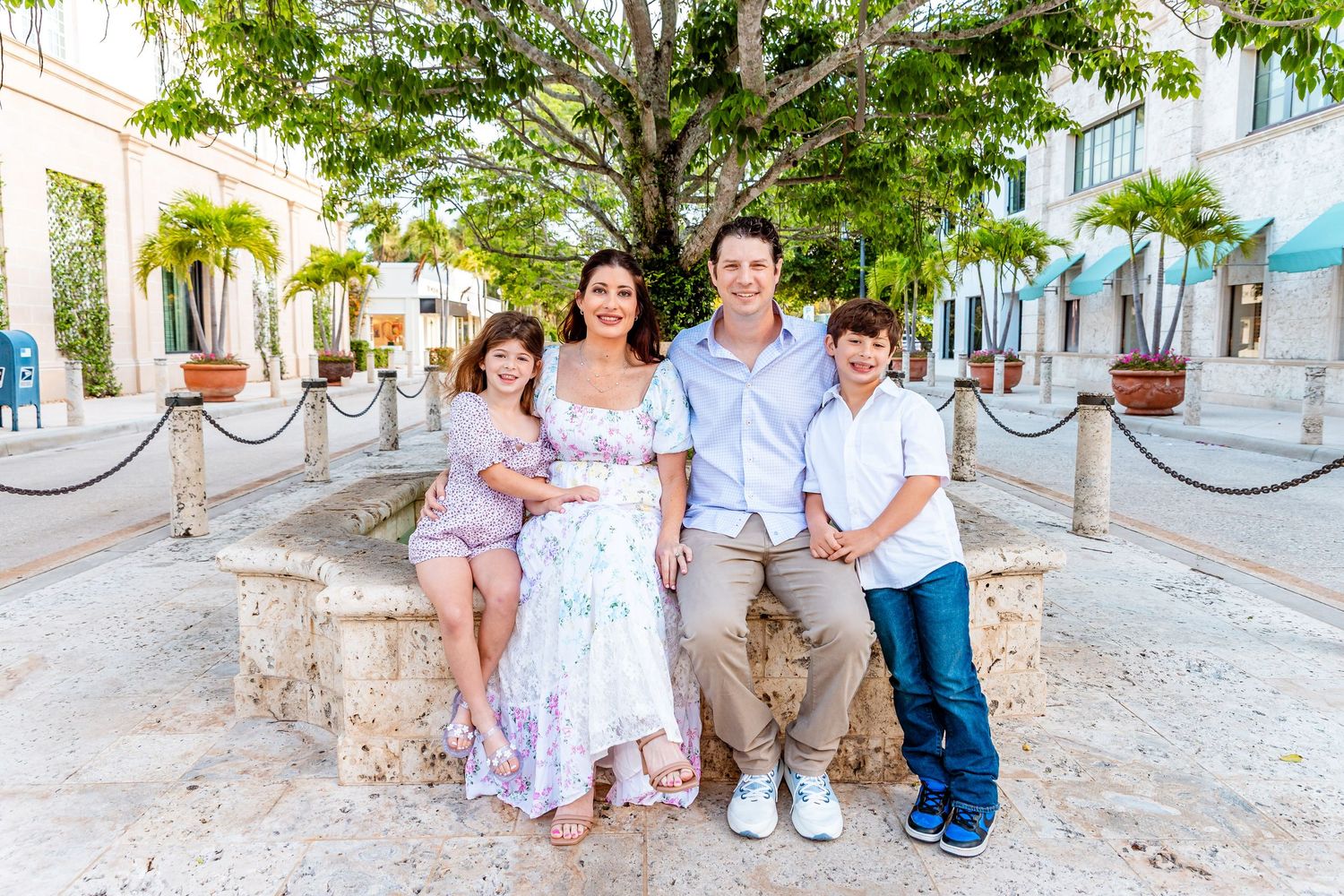 Family of four posing outdoors in a tropical setting, smiling together near a stone fountain.
