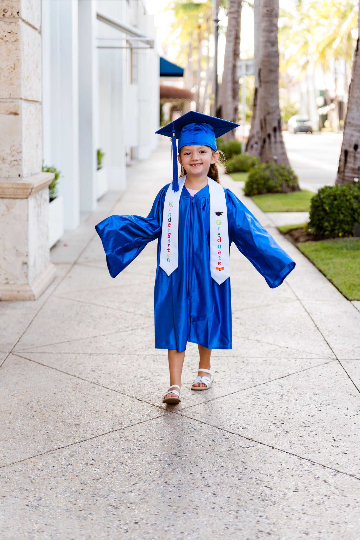 A child in blue graduation regalia walks under a white columned walkway, captured from multiple angles.