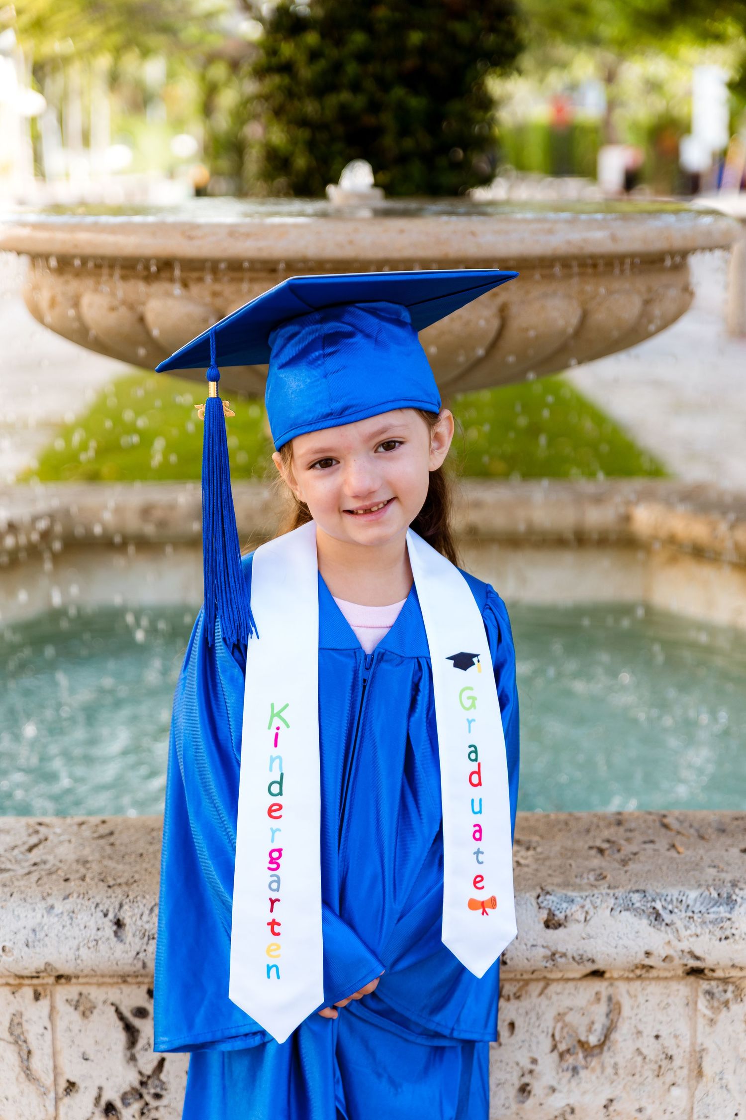 A young graduate in bright blue cap and gown stands smiling by a fountain wearing decorative kindergarten graduation stoles.