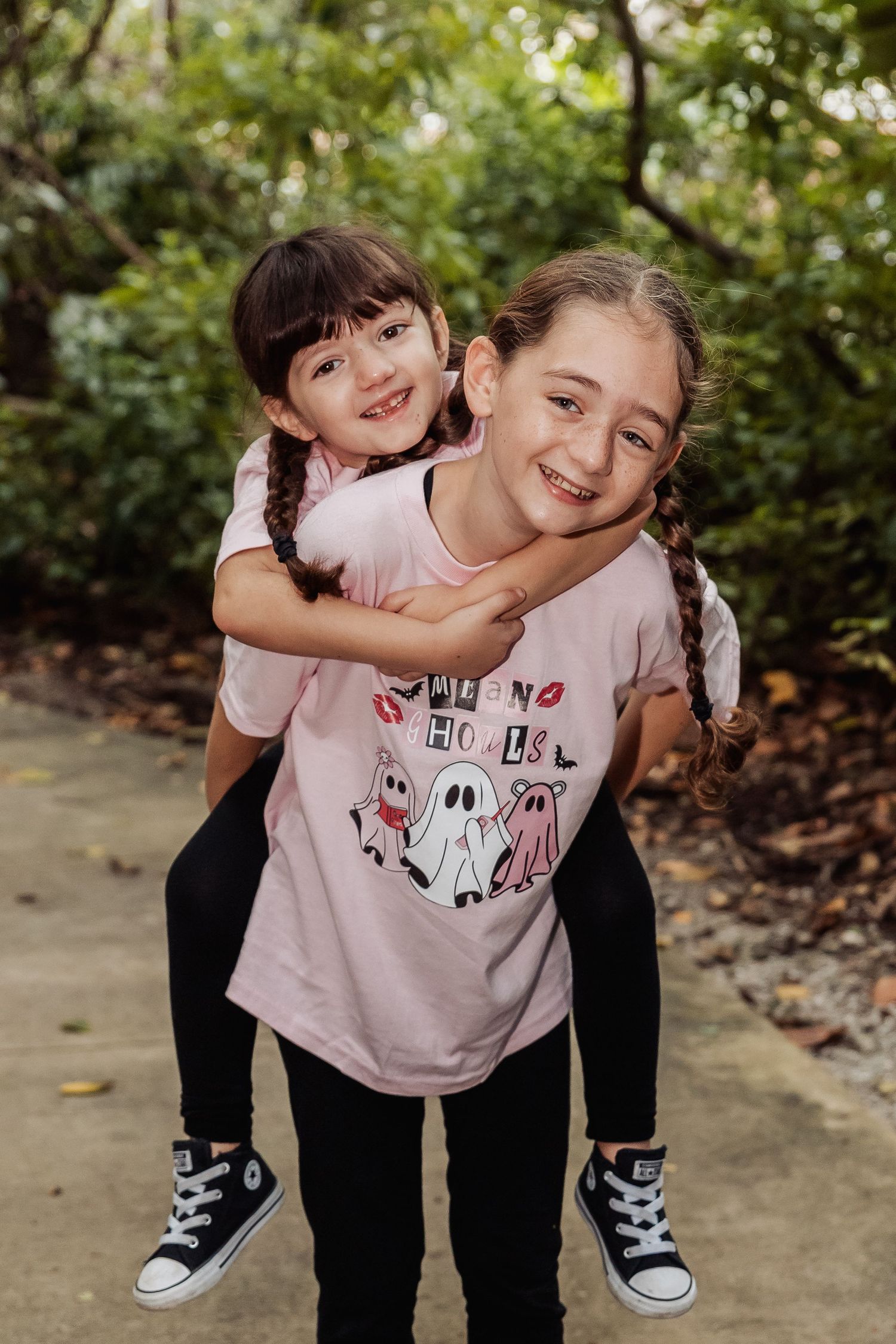 Two children in casual wear share a playful piggyback ride moment on a garden path surrounded by greenery.