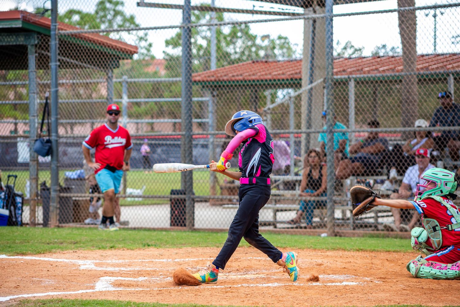 Youth softball player in purple uniform swings bat at home plate during game with spectators watching.